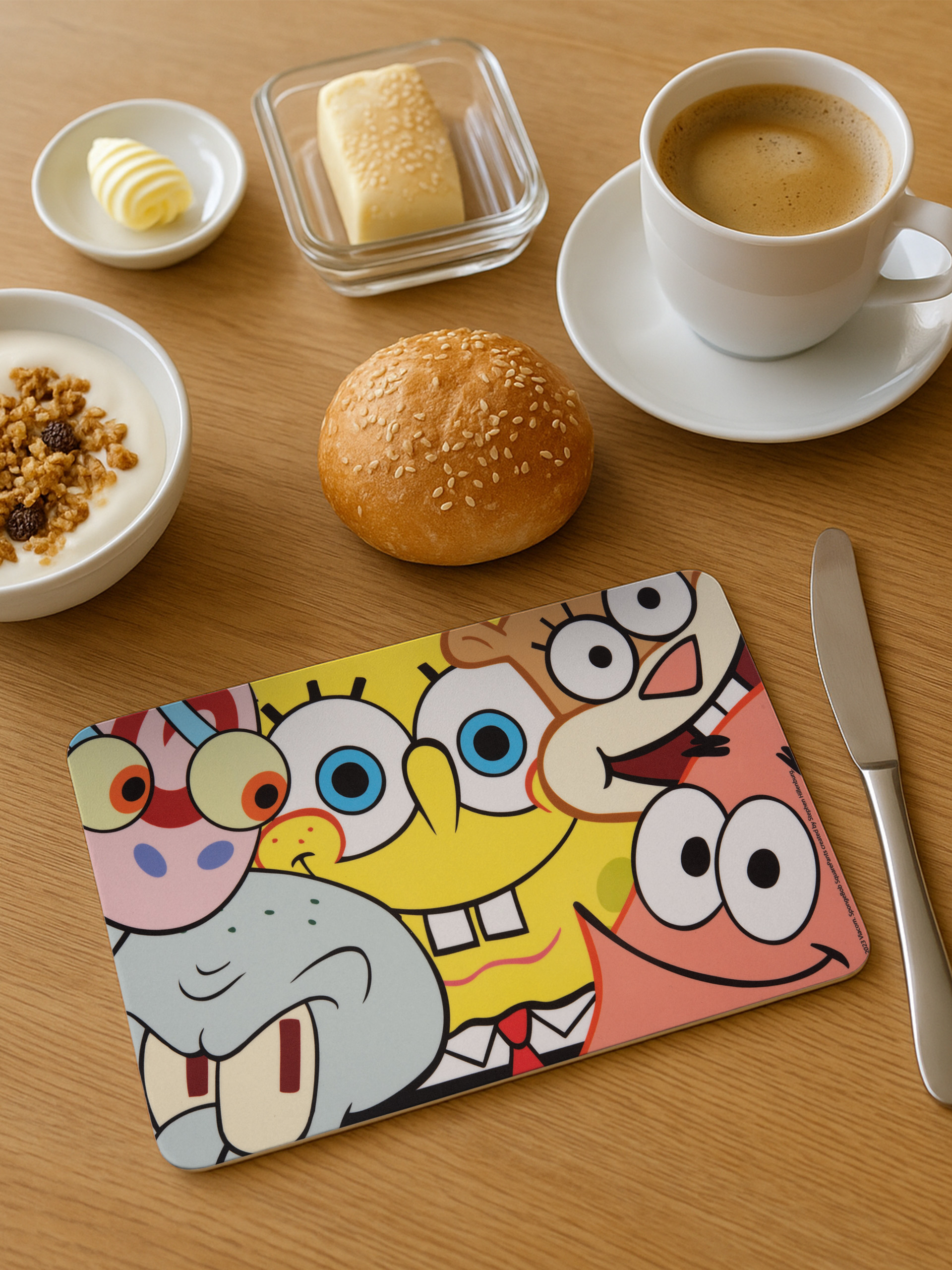 Frühstücksszene mit Kaffee, Joghurt, Butter, Brot und einer SpongeBob-Tischunterlage auf einem Holztisch.