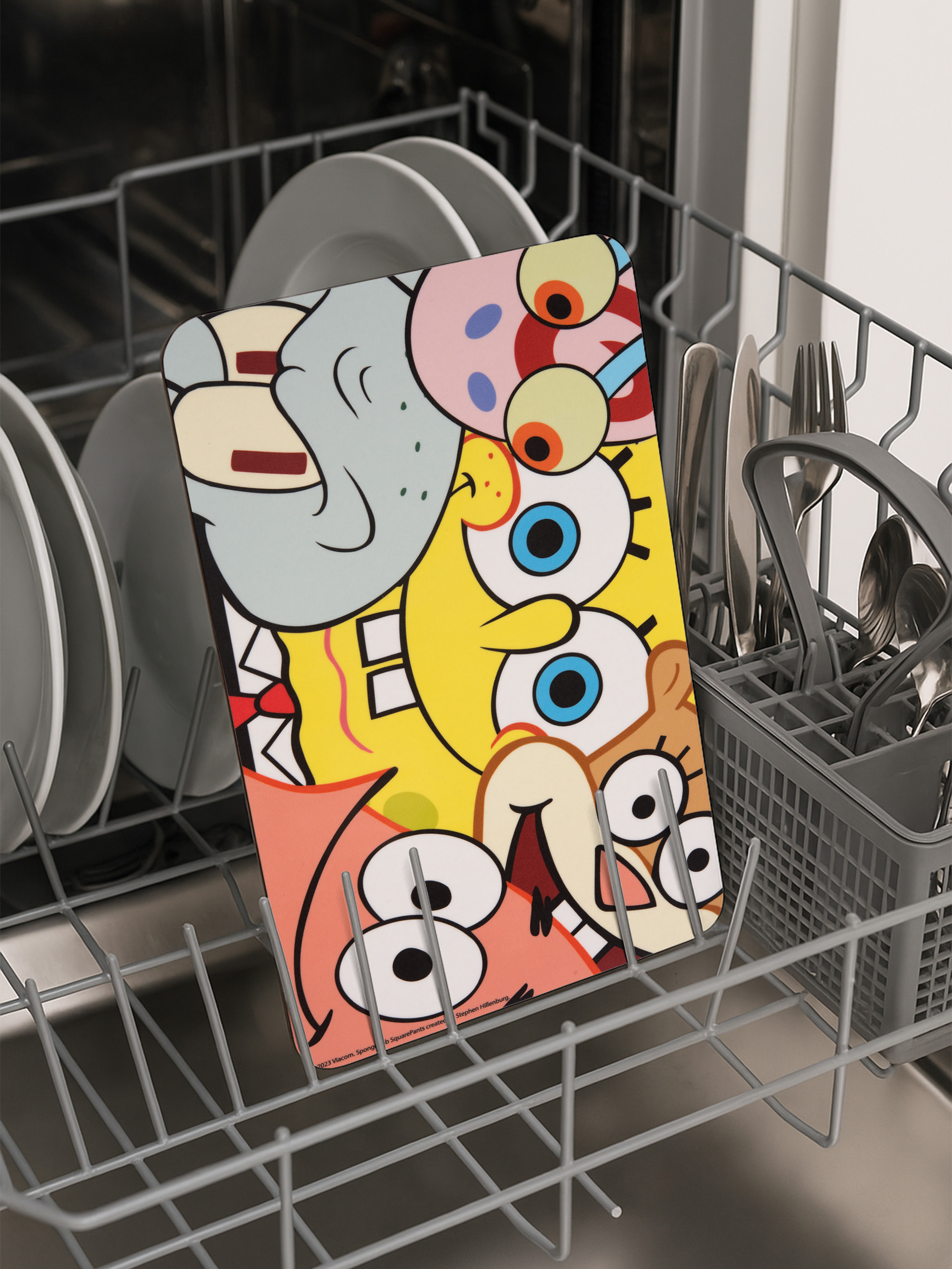 Ein buntes Spongebob Squarepants Schneidebrett befindet sich in einer Spülmaschine. Geschirr und Besteck sind ebenfalls vorhanden.