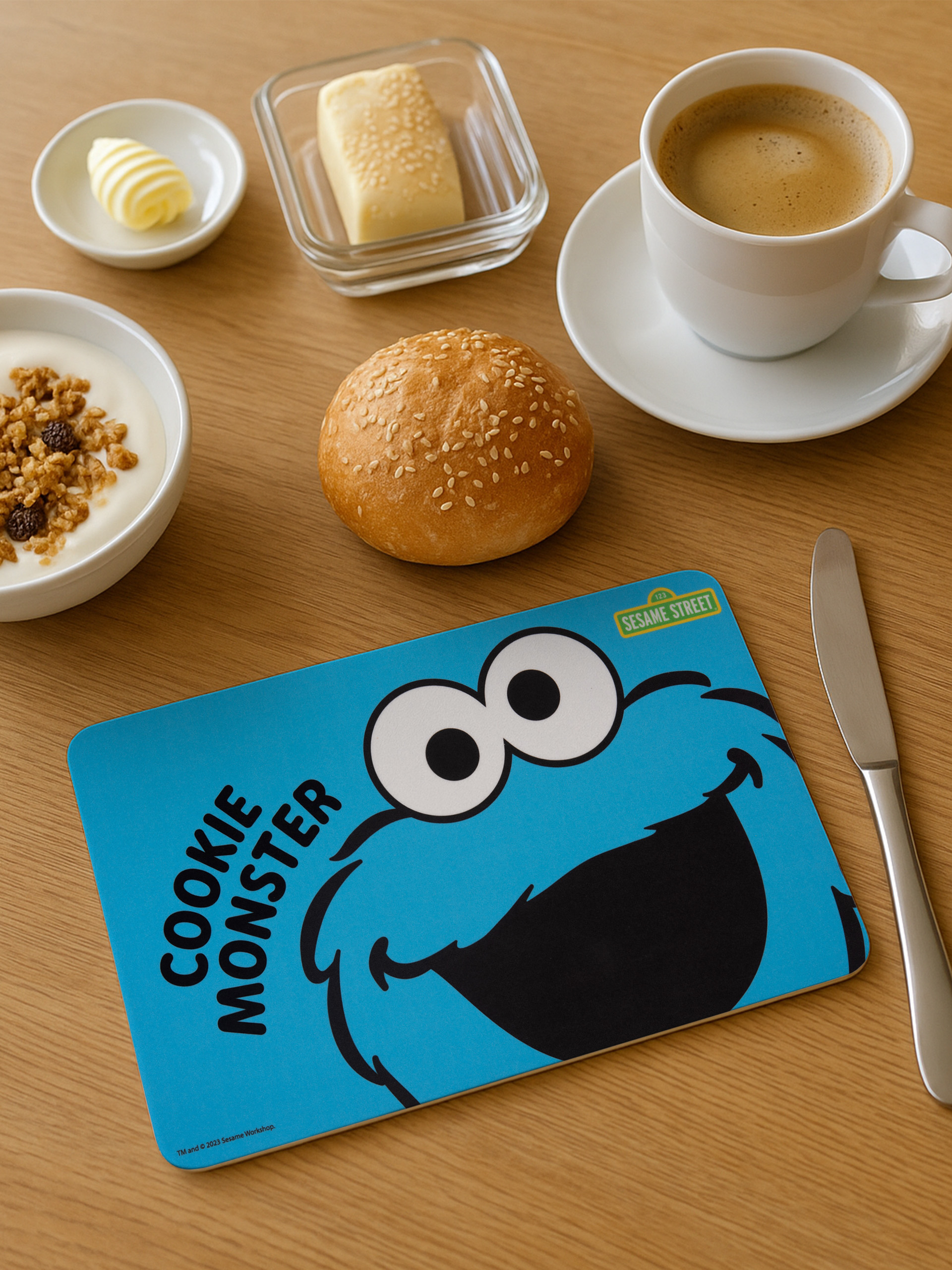Frühstücksszene mit Cookie Monster Platzset, Kaffee, Butter, Joghurt und Brötchen auf Holztisch.