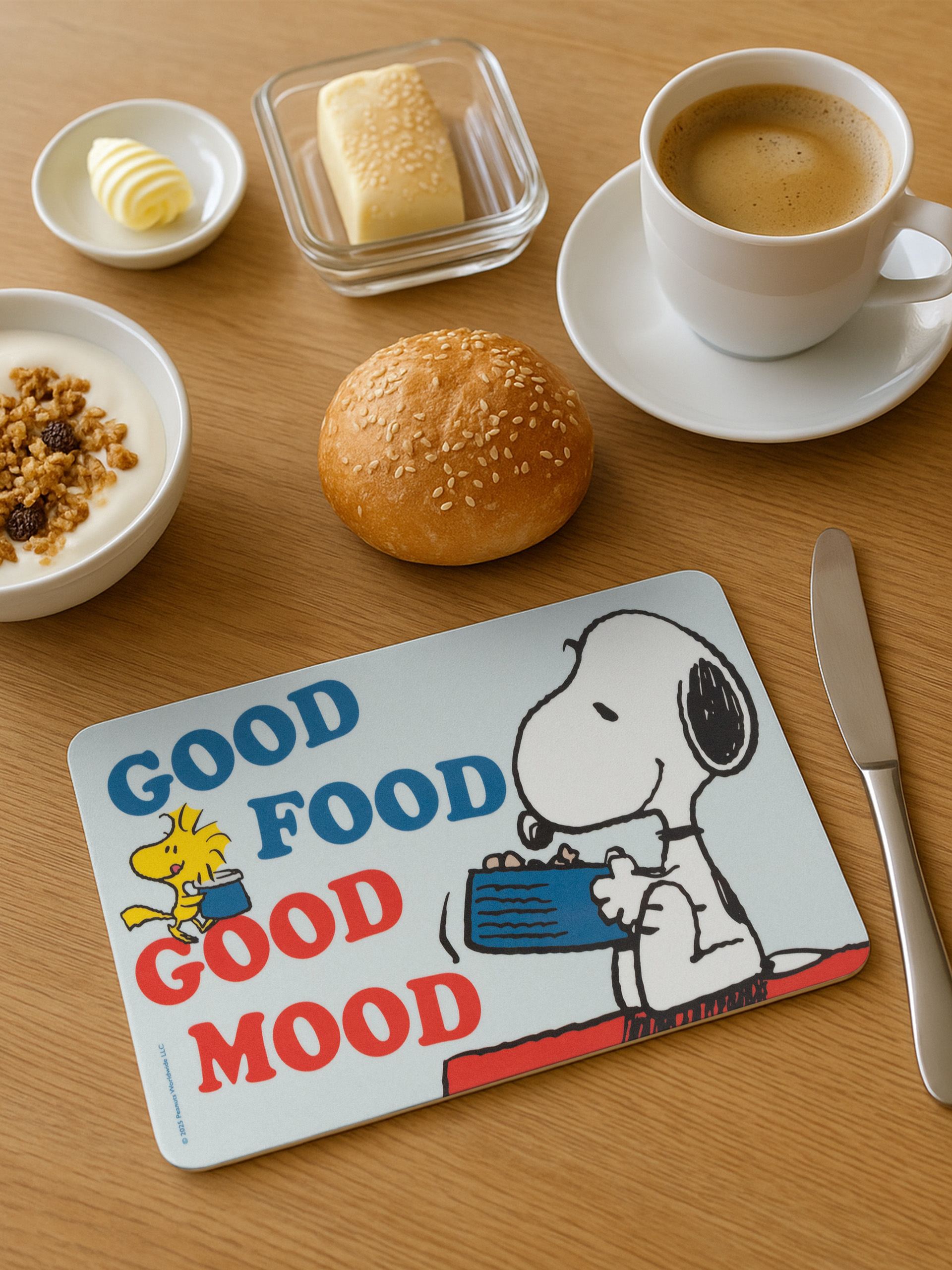 Frühstücksszene: Kaffee, Butter, Joghurt, Brötchen und eine Snoopy-Tischmatte mit 'Good Food Good Mood'.
