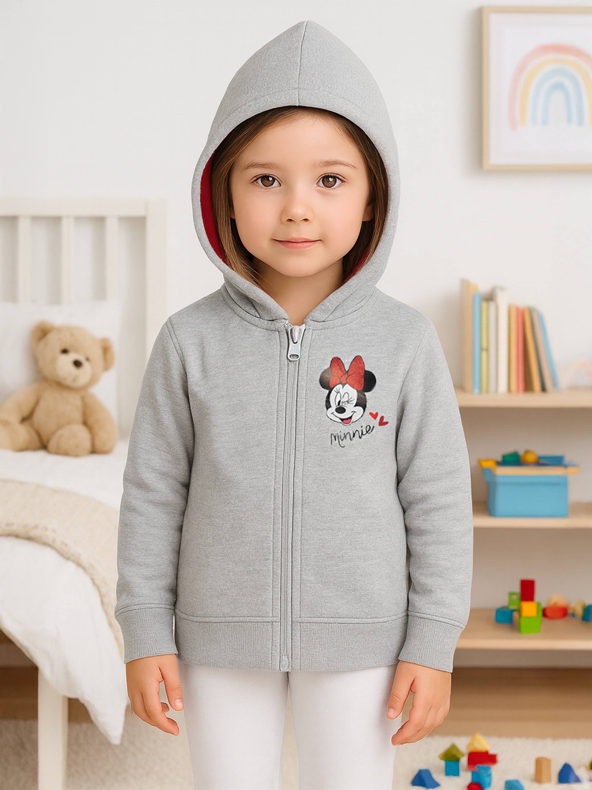 Ein Mädchen in einem grauen Minnie-Maus-Kapuzenpullover steht in einem Zimmer. Ein Teddybär und ein Bücherregal sind sichtbar.