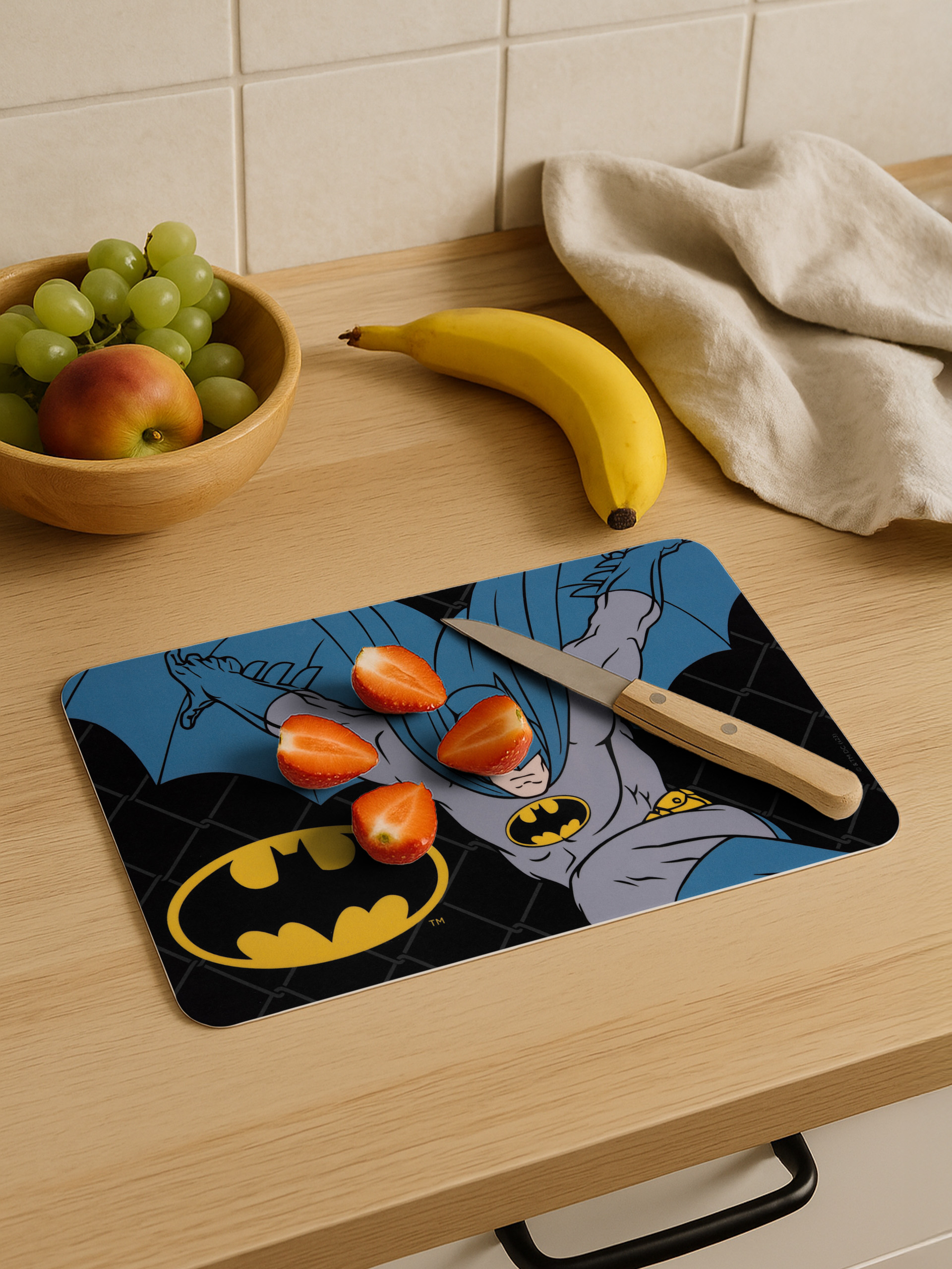 Ein Schneidebrett mit Batman-Logo, Erdbeeren, Banane, Trauben und Apfel auf einem Holztisch.
