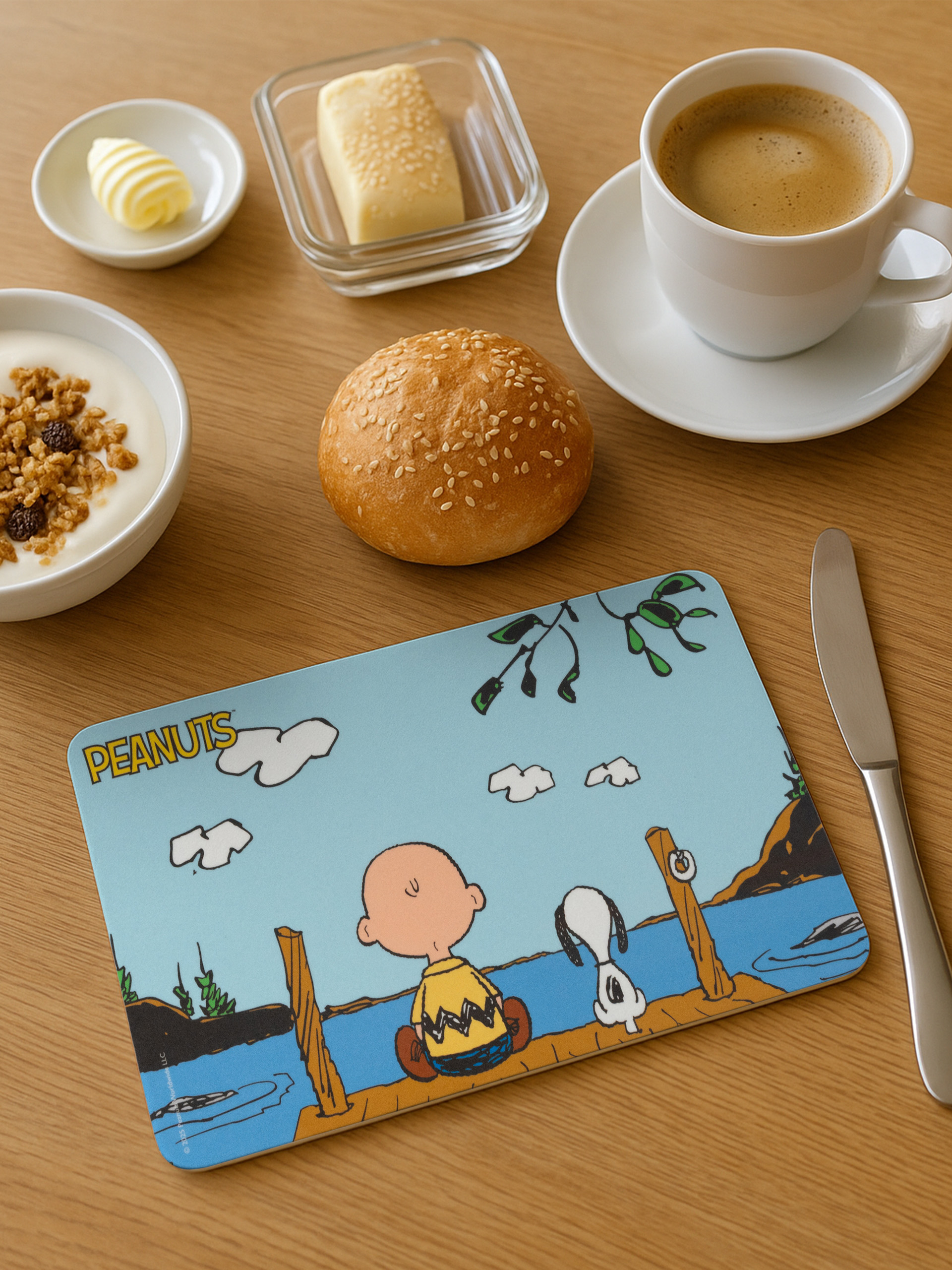 Frühstücksszene: Kaffee, Butter, Joghurt, Brötchen, Peanuts-Tischset mit Charlie Brown und Snoopy auf einem Steg.
