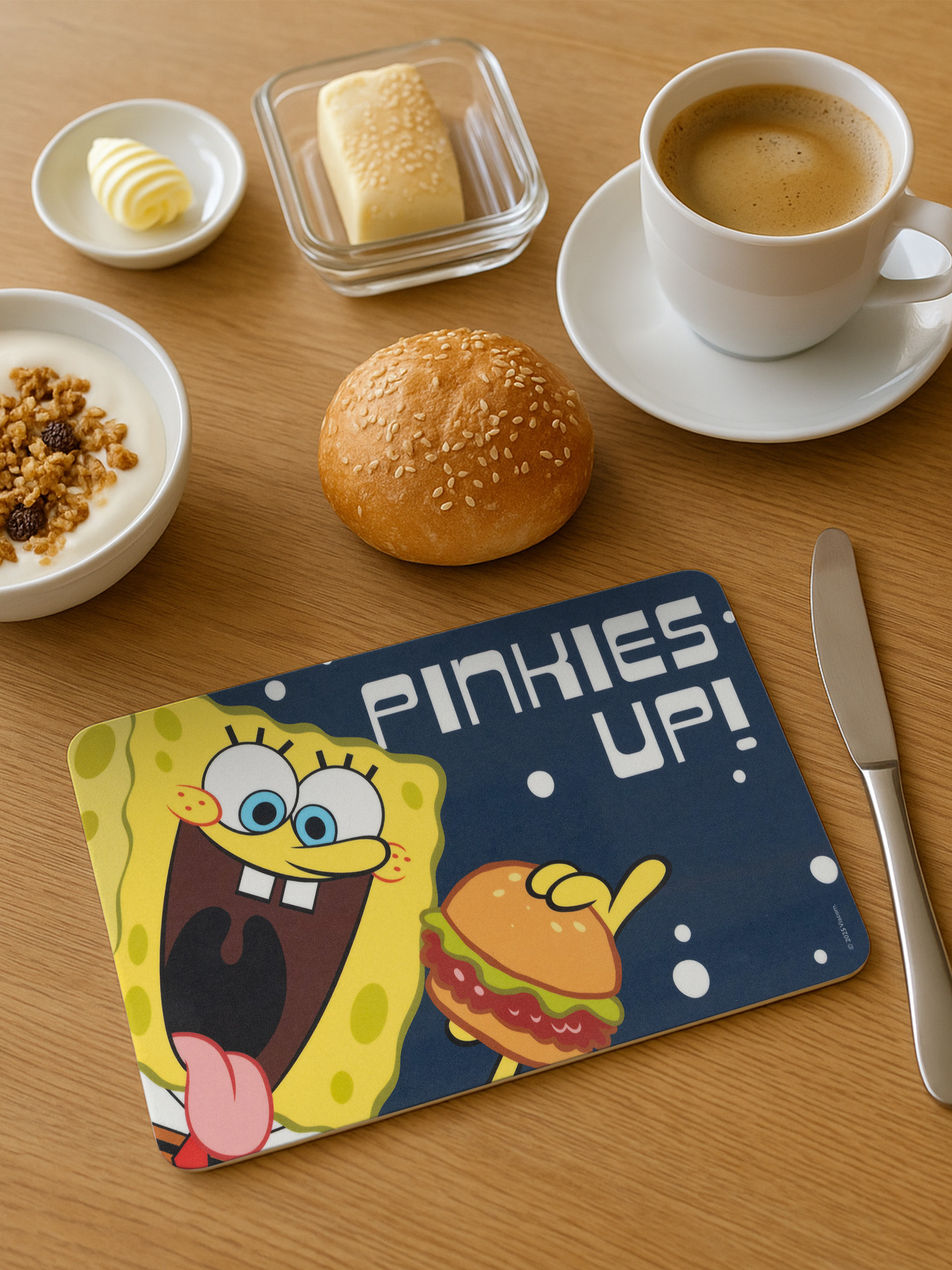 Frühstücksszene mit Kaffee, Joghurt, Butter, Brot und einer SpongeBob-Tischdecke mit Burger.