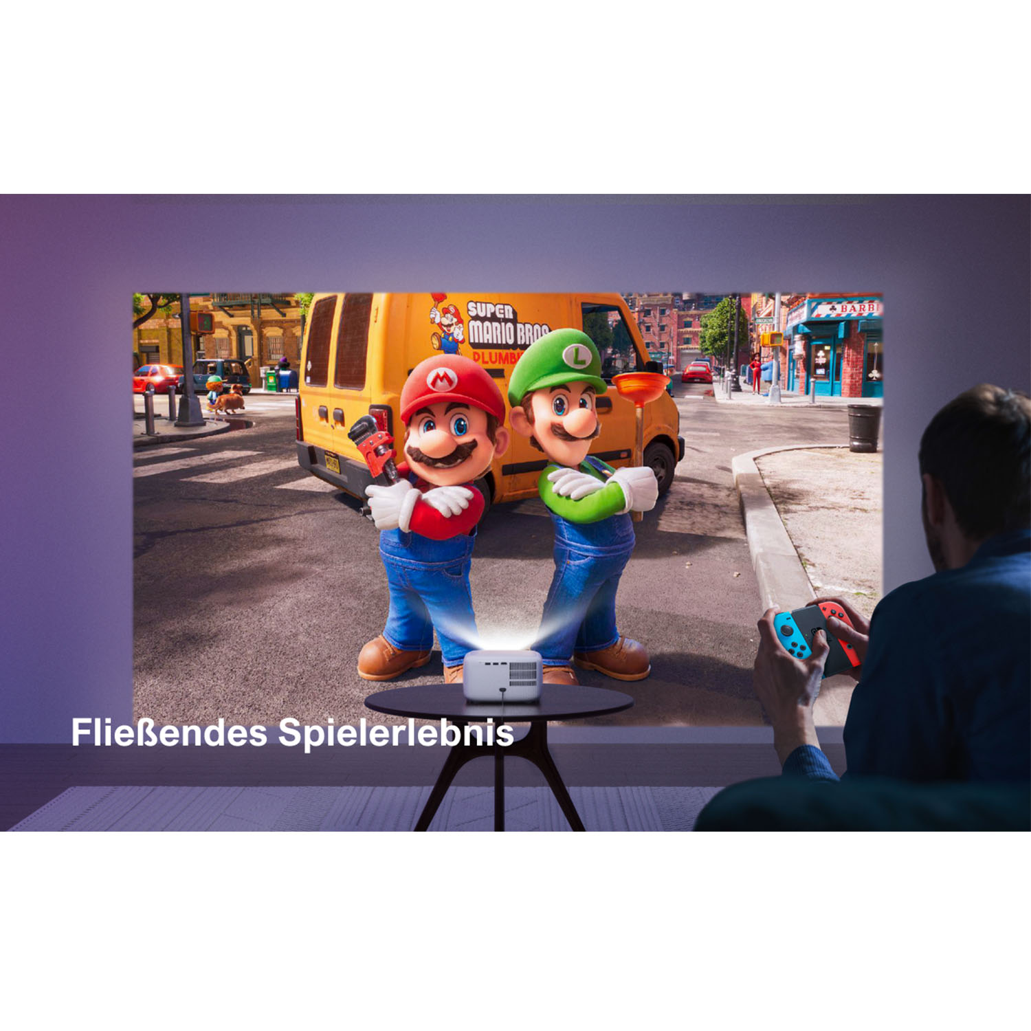 Mario und Luigi auf einem Bildschirm, projiziert von einem Gerät. Eine Person hält einen Game-Controller. Der Hintergrund zeigt einen gelben Van.