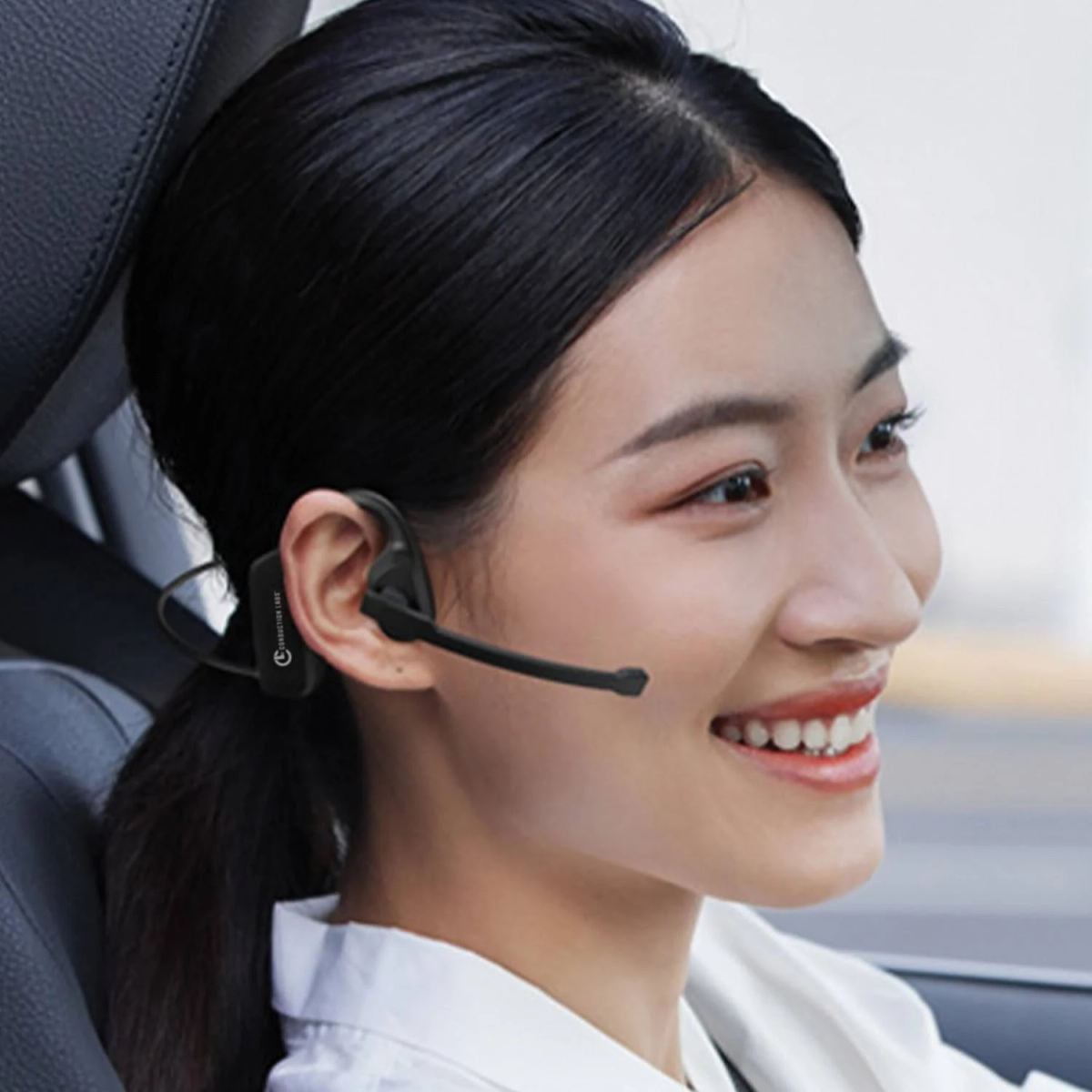 Una donna con auricolare nero sorride in auto. Ha i capelli neri legati e indossa una camicia bianca.
