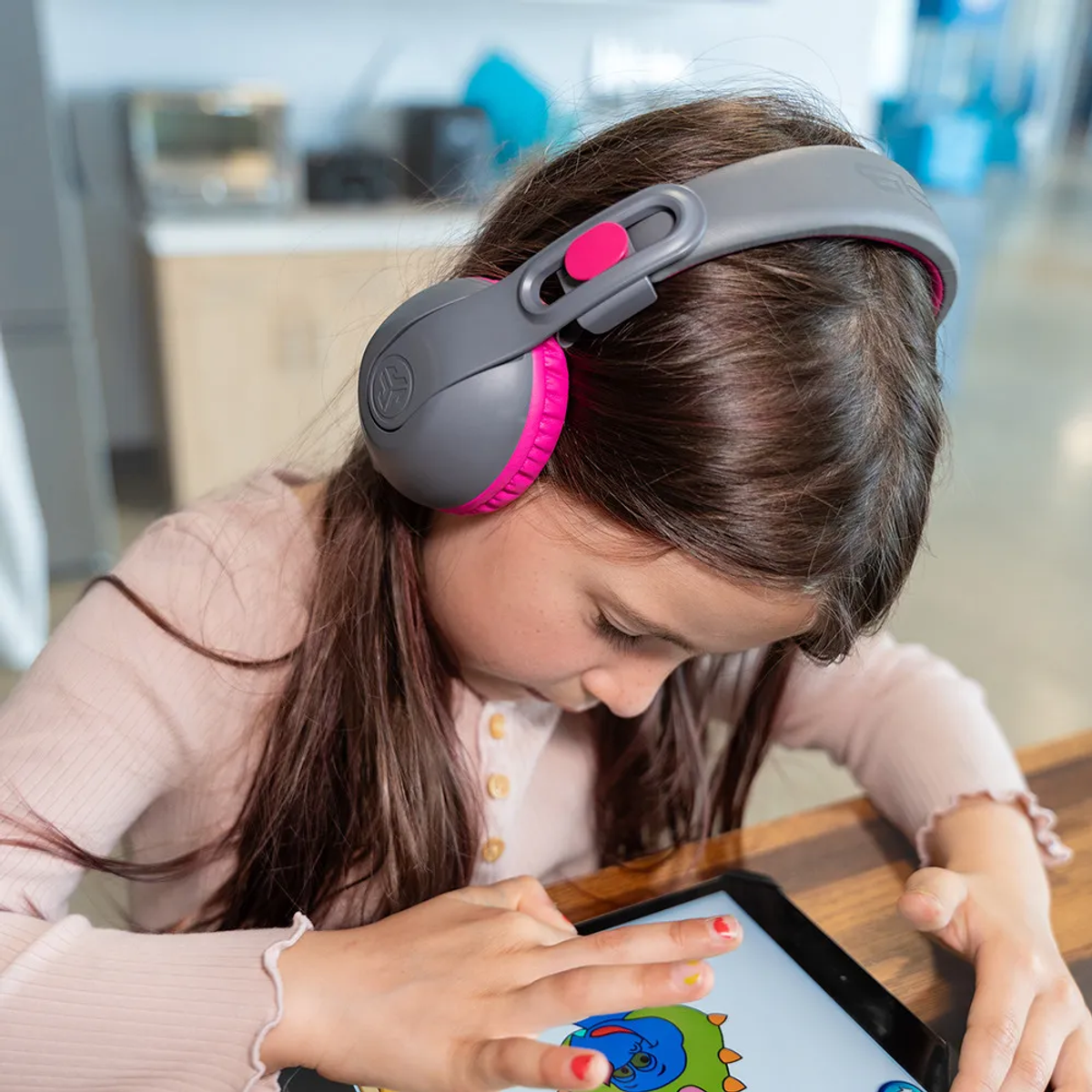 Una ragazza con le cuffie usa un tablet. Le cuffie sono grigie e rosa. La ragazza indossa una camicia rosa e il tablet mostra un cartone animato.