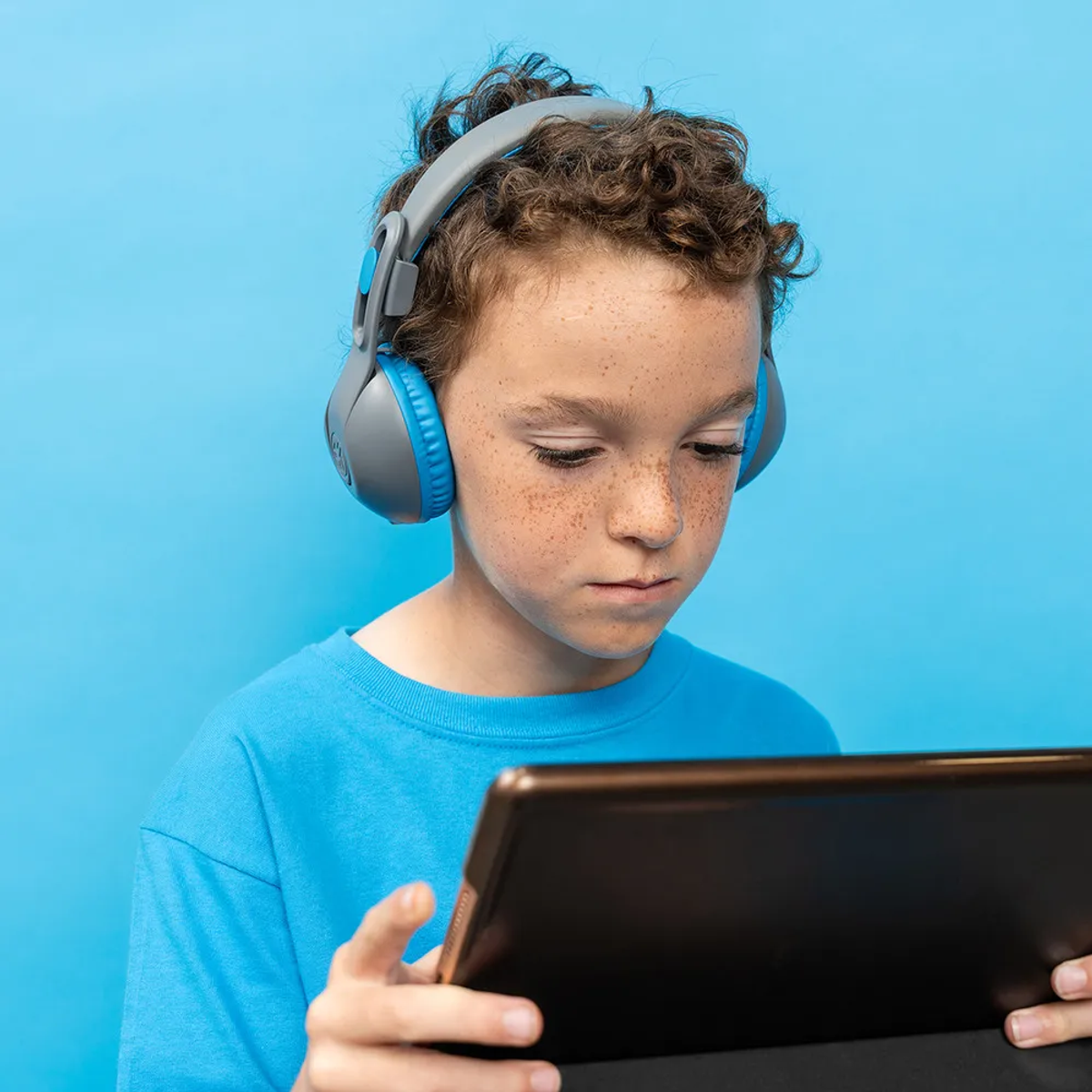 Un ragazzo con i capelli ricci e lentiggini, con le cuffie, guarda un tablet. Lo sfondo è blu.