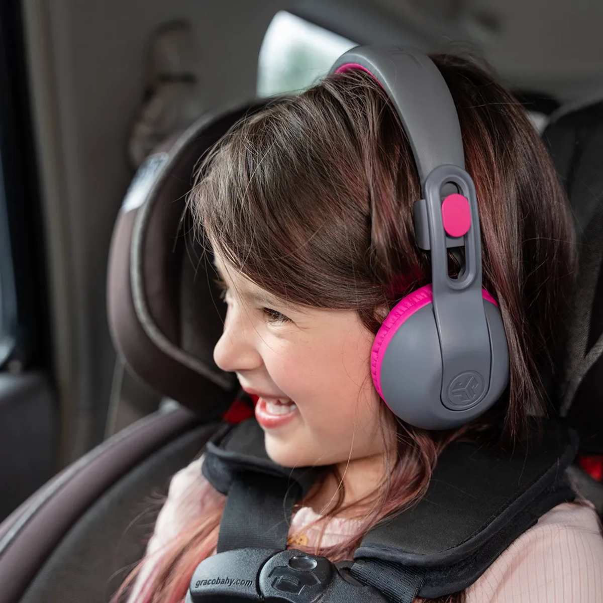 Una ragazza sorride con cuffie grigie e rosa in un seggiolino. È allacciata e guarda di lato.