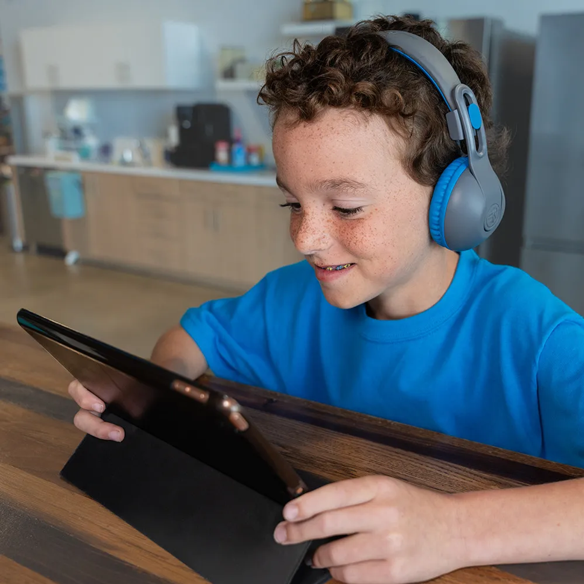 Un ragazzo con le cuffie guarda un tablet. Indossa una maglietta blu, seduto a un tavolo di legno, con una cucina sullo sfondo.