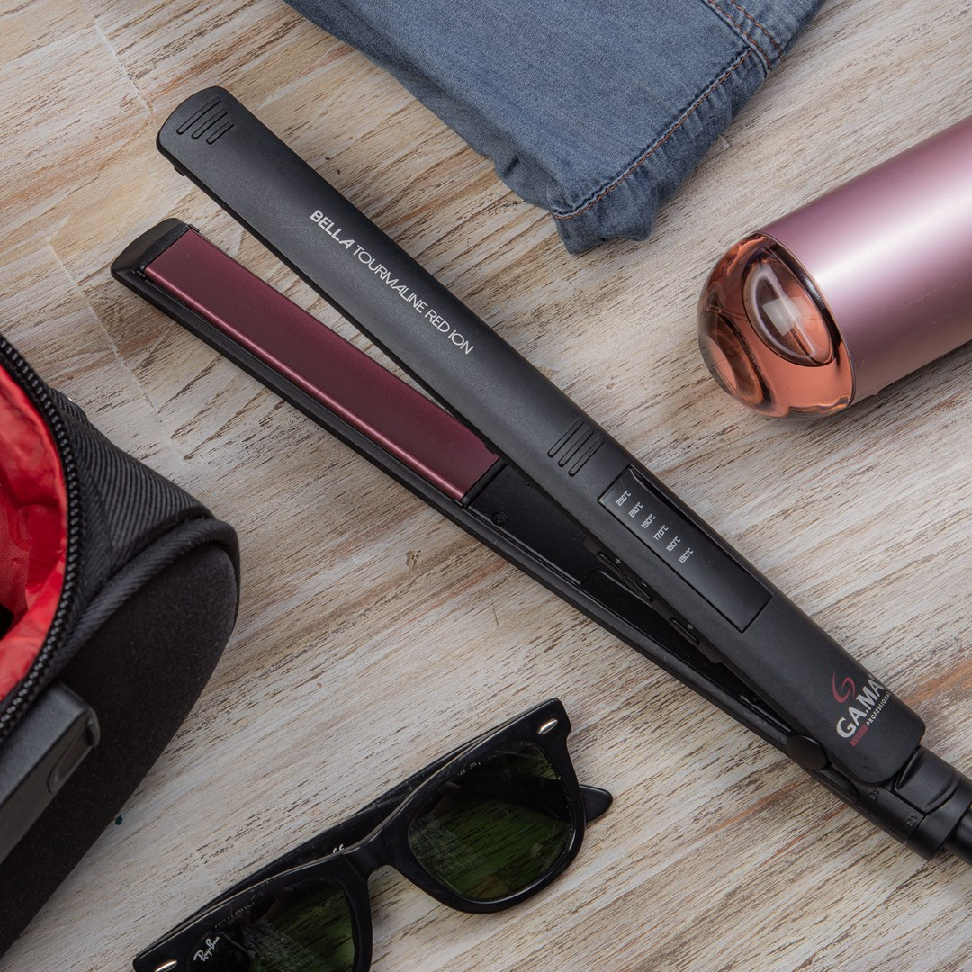 Una piastra per capelli, occhiali da sole, jeans, un beauty case e una bottiglia sono posizionati su una superficie di legno.