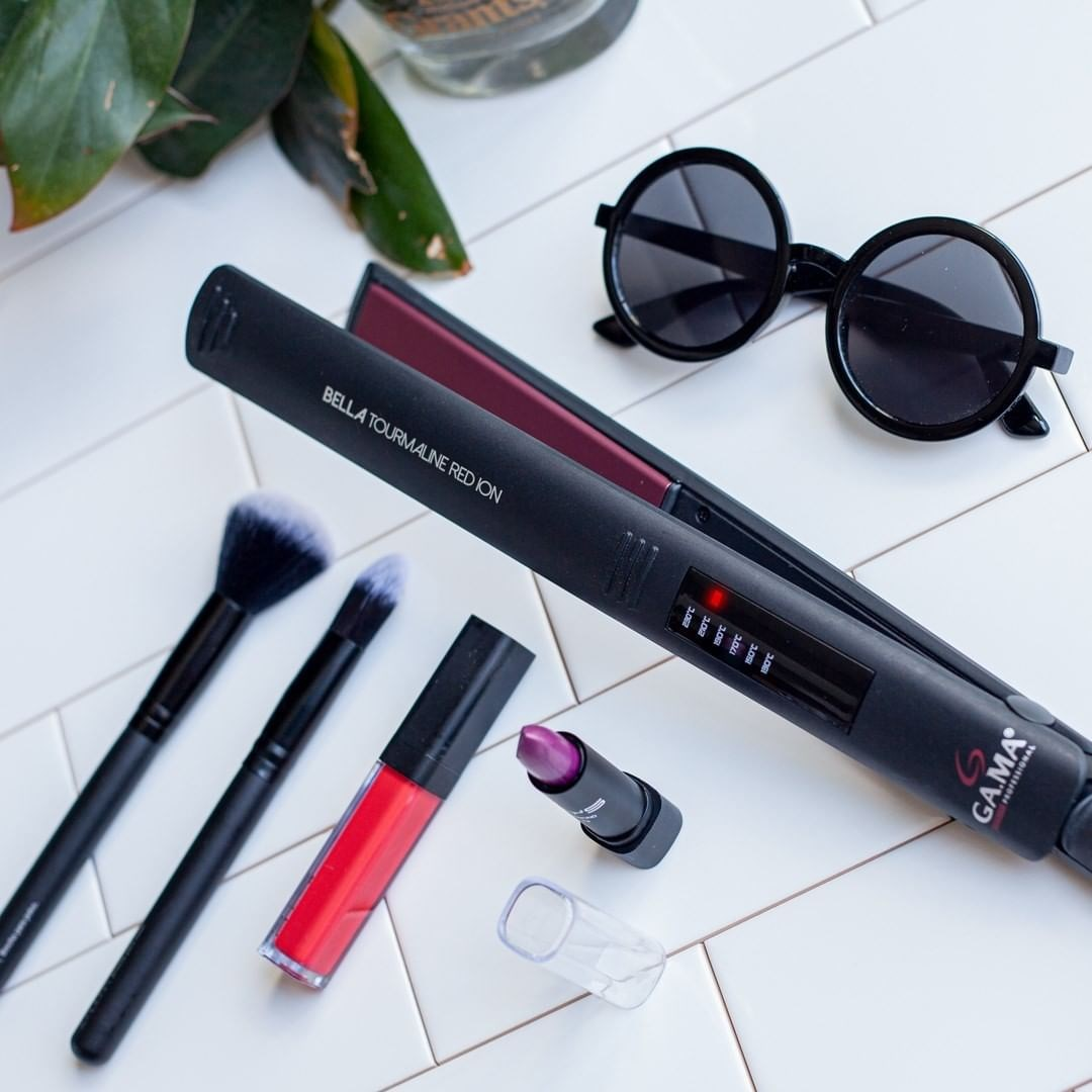 Piastra per capelli nera, occhiali da sole, pennelli per trucco, lucidalabbra rosso e rossetto su superficie piastrellata bianca.