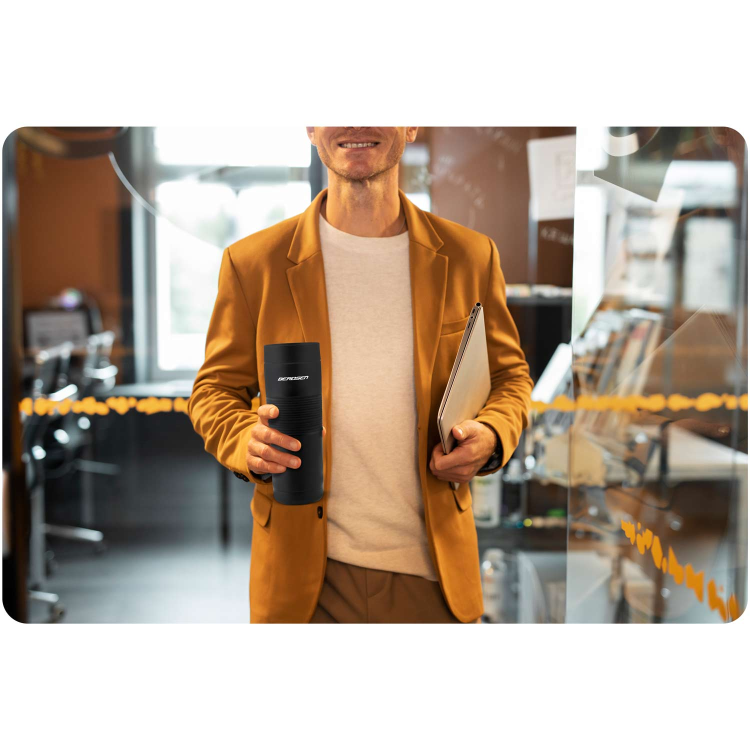 Mężczyzna w musztardowej marynarce trzyma czarny termos i laptopa. Uśmiecha się w otoczeniu biurowym.