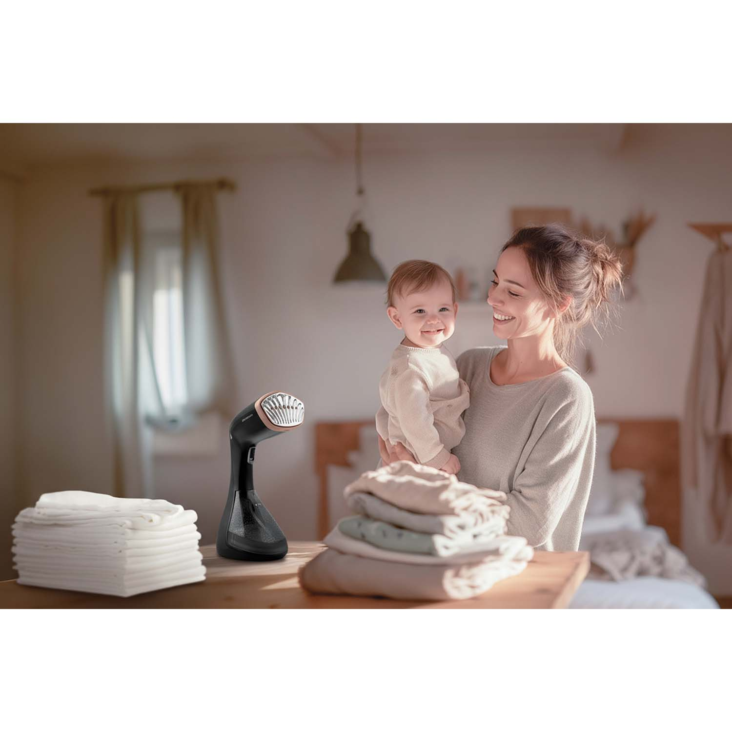 Kobieta trzyma uśmiechnięte dziecko. Parownica do ubrań stoi na stole w pobliżu złożonych ubrań i stosu białych ręczników.