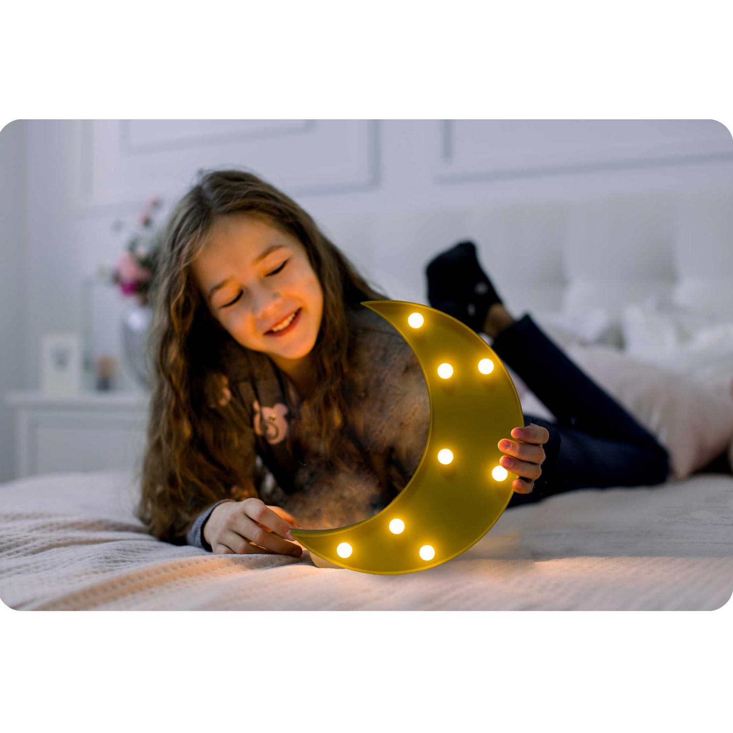 Dziewczyna uśmiecha się trzymając podświetlaną lampę w kształcie półksiężyca na łóżku. Scena jest delikatnie oświetlona, z przytulną atmosferą.