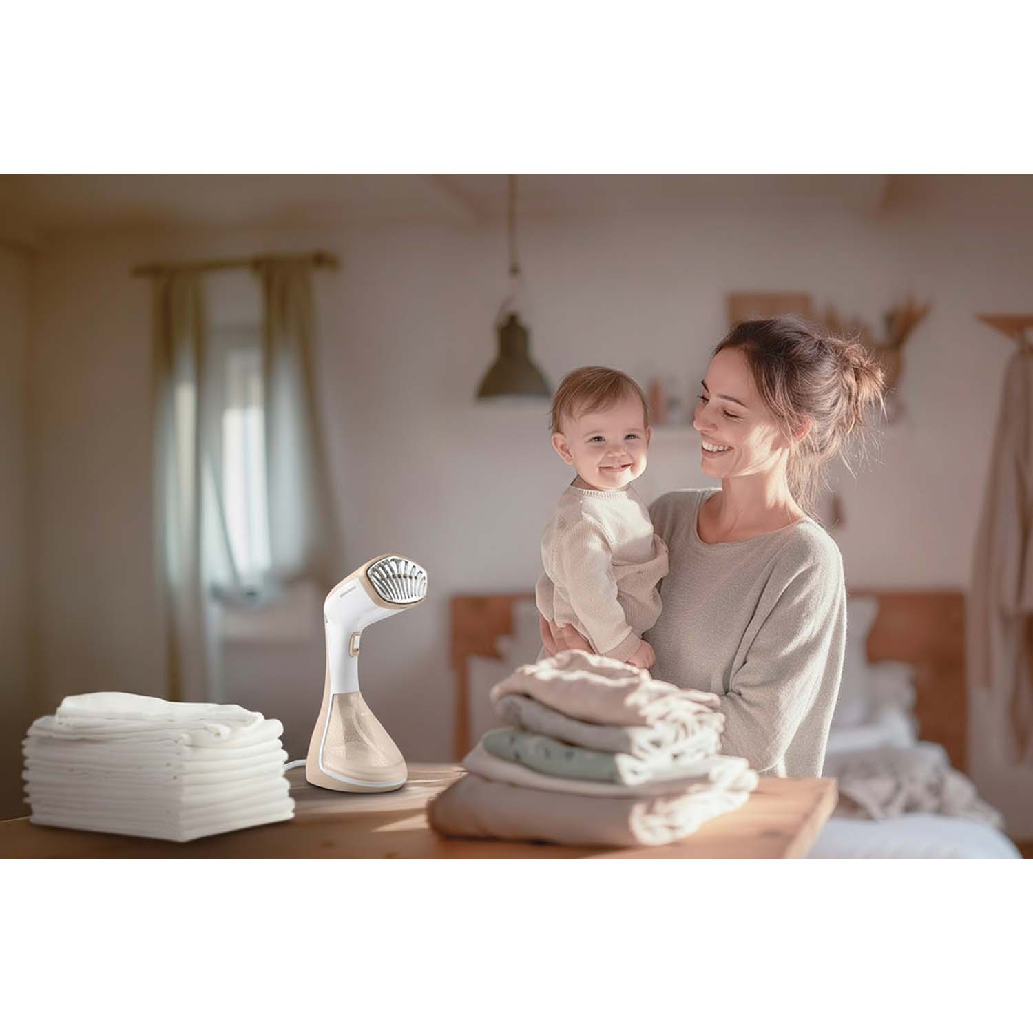 Kobieta trzyma dziecko, uśmiechając się. Parownica do ubrań na stole ze złożonymi ubraniami. Ciepła, domowa scena.