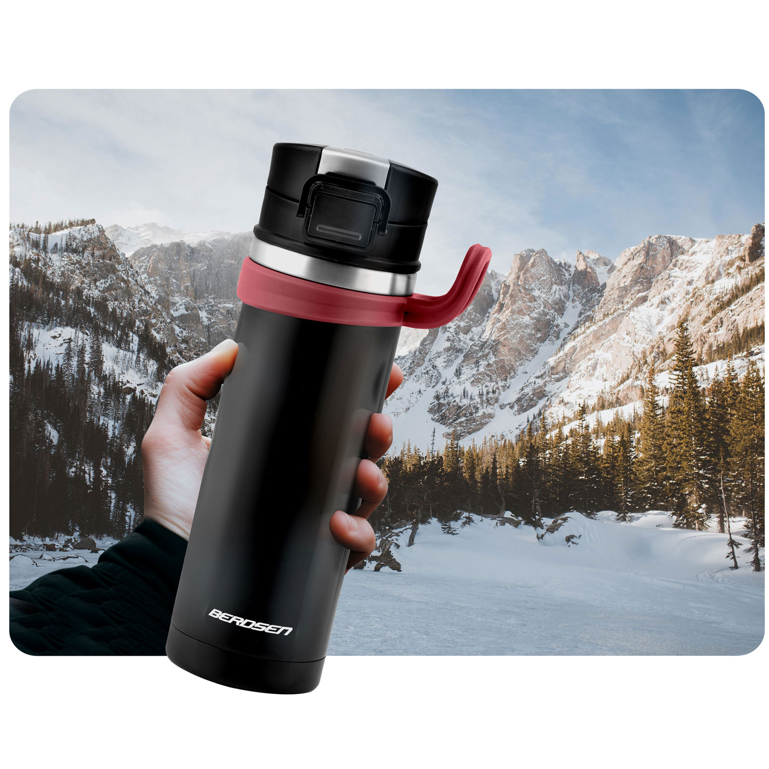 Ręka trzyma czarny termos z czerwonym uchwytem. W tle góry ze śniegiem i drzewami pod błękitnym niebem.