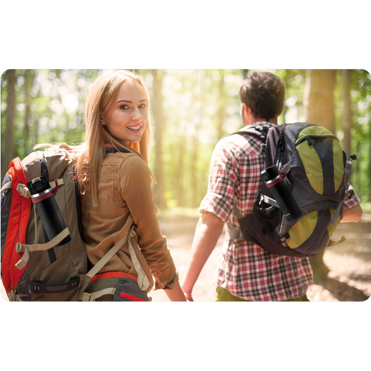 Para wędruje po lesie, trzymając się za ręce. Mają plecaki z butelkami wody. Kobieta uśmiecha się do widza.