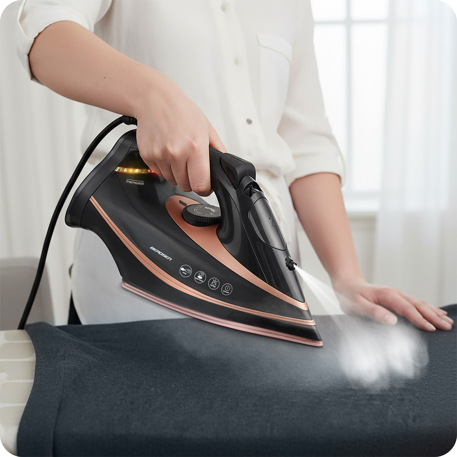 Osoba prasuje ciemny element garderoby czarnym i różowo-złotym żelazkiem, unosi się para. Osoba ma na sobie białą koszulę.