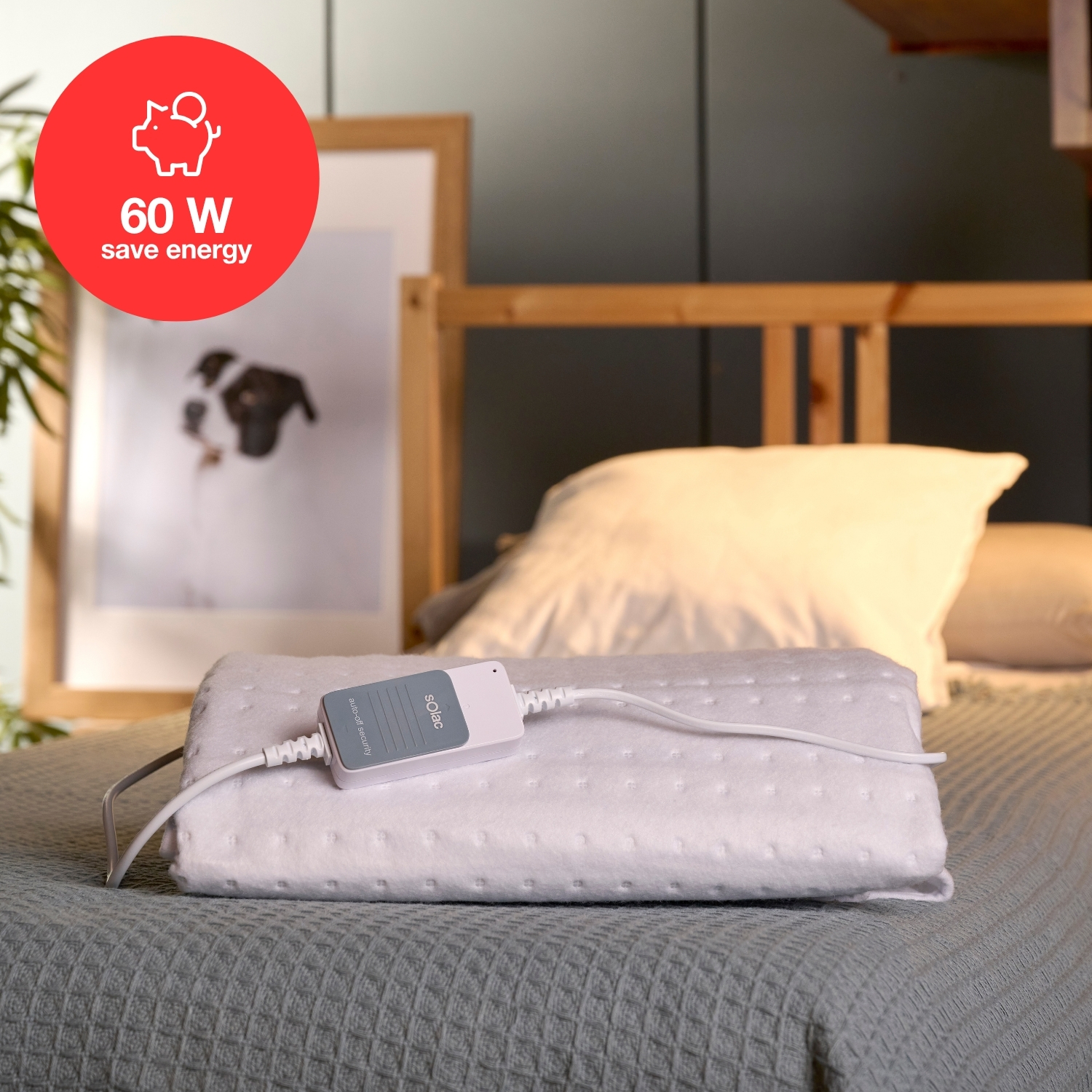 Biały koc elektryczny z panelem sterowania leży na łóżku. W tle obraz w ramie i znak oszczędzania energii 60W.