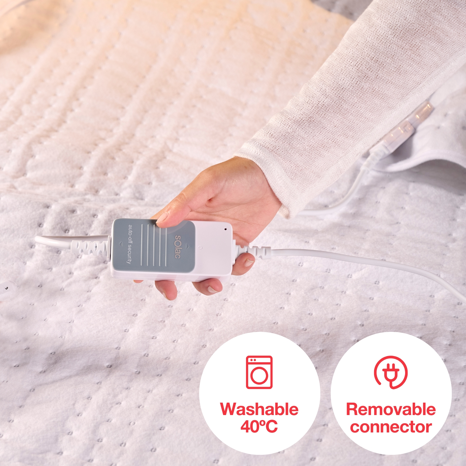 Ręka trzyma biało-szare urządzenie Solac na białym kocu. Ikony wskazują, że można je prać w 40°C i ma odłączany konektor.