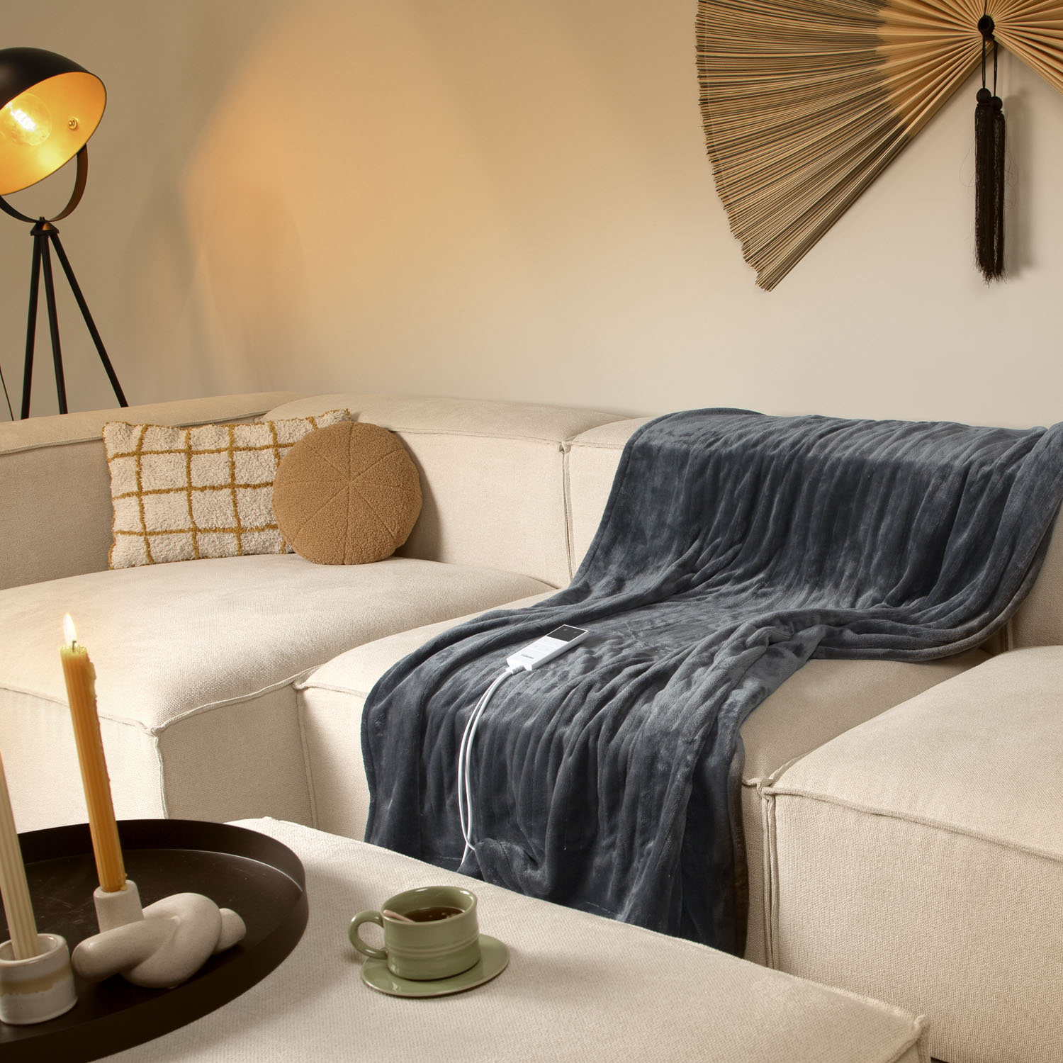 A gray blanket on a beige sofa, a lit candle, a coffee cup, and a decorative fan on the wall are visible.