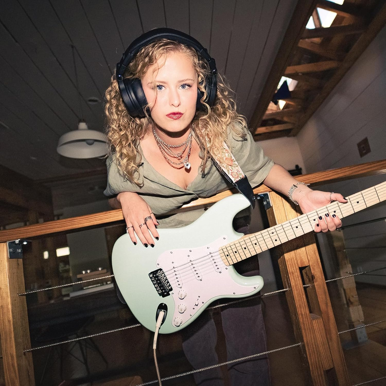 Vrouw met koptelefoon houdt een mintgroene gitaar vast, leunend op een houten reling. Ze draagt ringen en een ketting en kijkt de camera in.