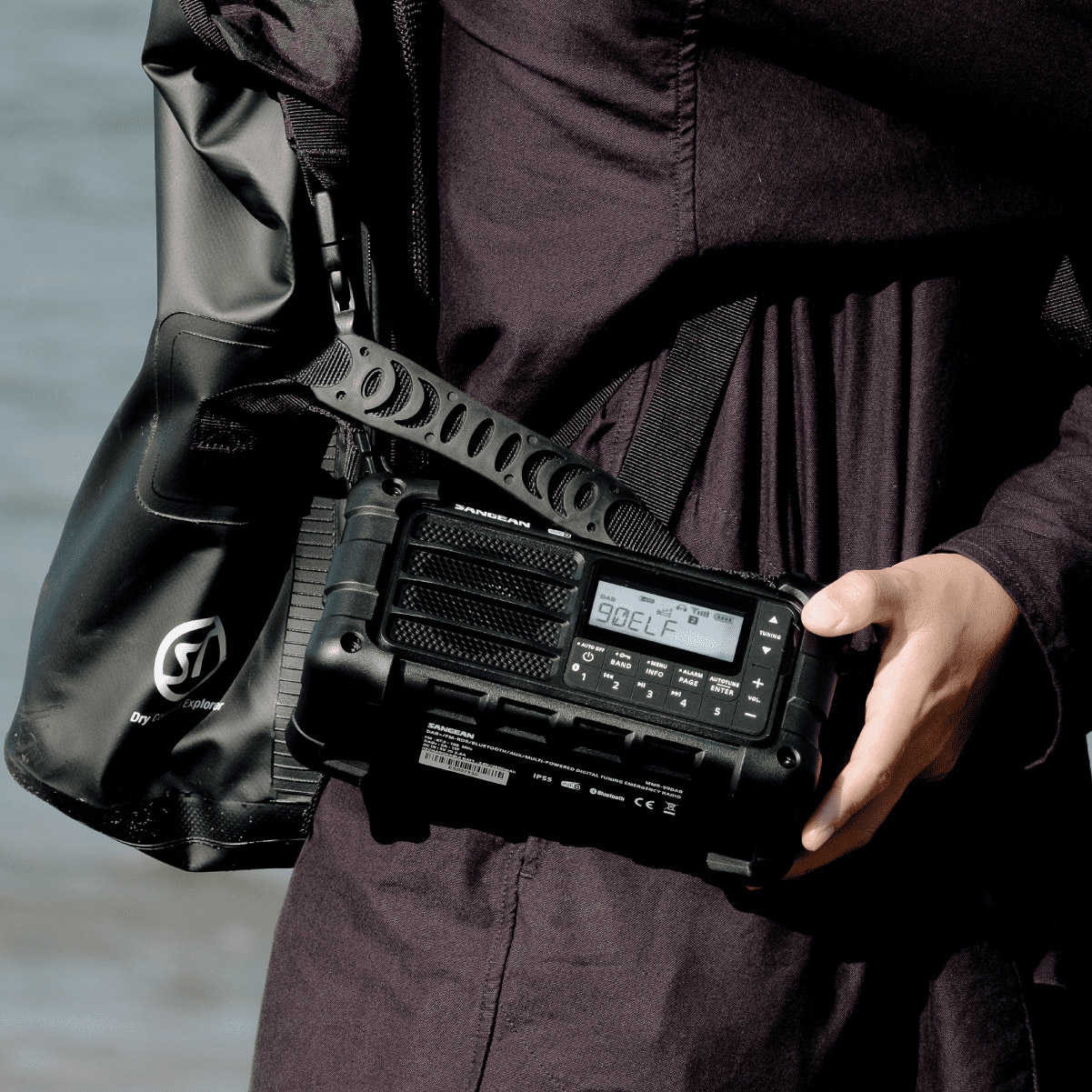 Eine Person hält ein schwarzes Sangean-Radio. Eine schwarze Tasche hängt an ihrer Schulter. Der Hintergrund ist eine verschwommene Wasserszene.