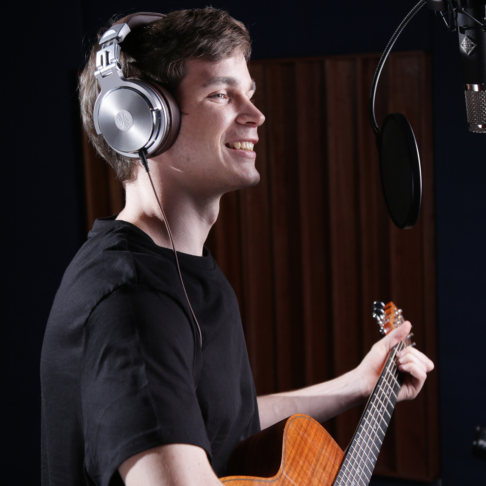 Un uomo con le cuffie e la chitarra sorride in uno studio di registrazione davanti a un microfono.