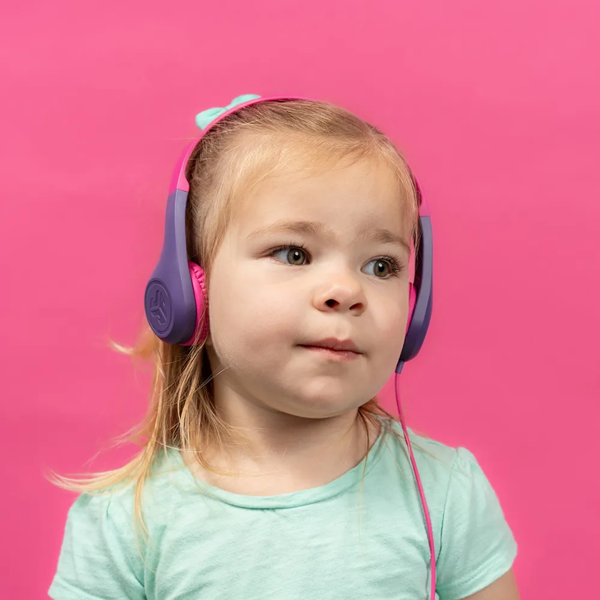 Mała dziewczynka nosi różowe i fioletowe słuchawki, patrząc lekko w bok. Ma blond włosy i nosi jasnozieloną koszulę, na różowym tle.