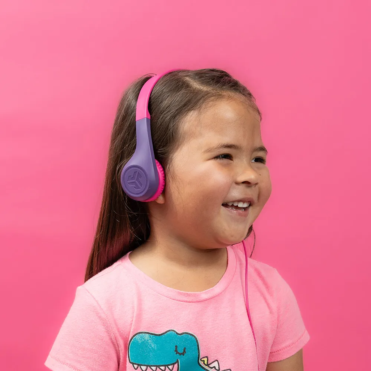 Uśmiechnięta dziewczynka w różowo-fioletowych słuchawkach. Ma różową koszulkę z obrazkiem dinozaura na różowym tle.