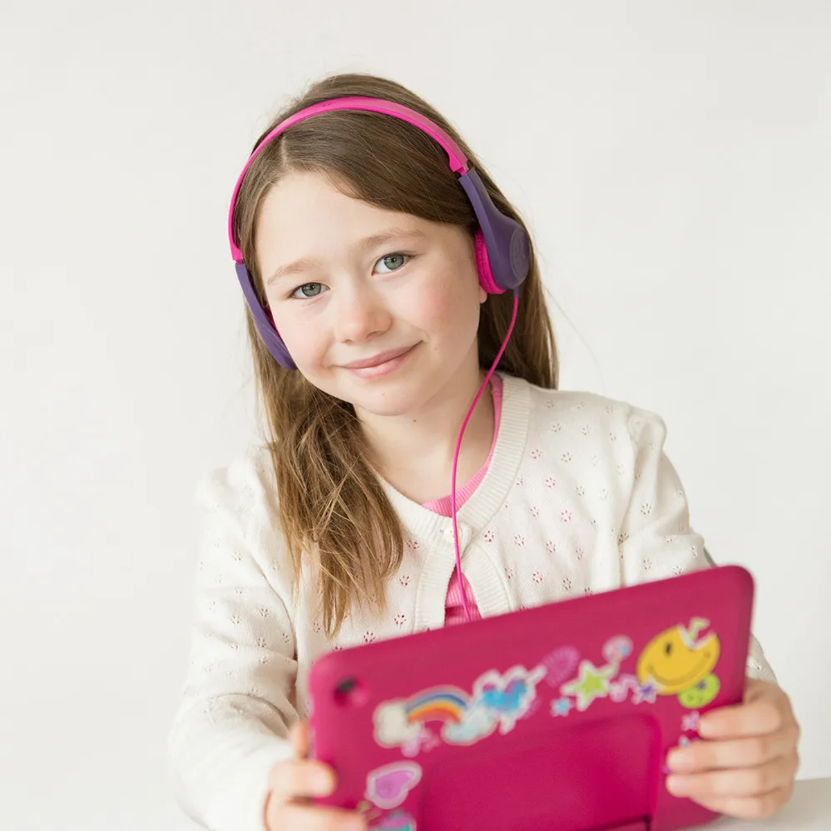 Dziewczyna w różowych słuchawkach trzyma różowy tablet. Ma biały sweter. Tablet ma naklejki.