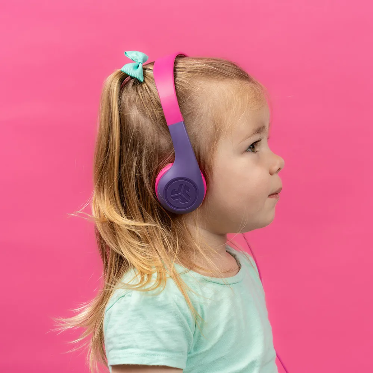 Mała dziewczynka nosi fioletowe i różowe słuchawki. Ma na sobie turkusową koszulkę i turkusową kokardę we włosach na różowym tle.