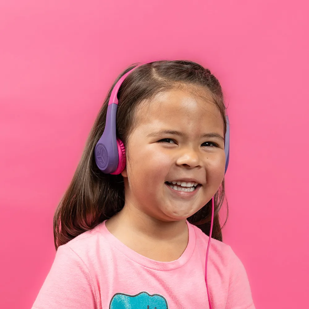 Uśmiechnięta dziewczynka w różowych słuchawkach. Ma na sobie różową koszulę, a tło jest również różowe. Słuchawki są fioletowe i różowe.