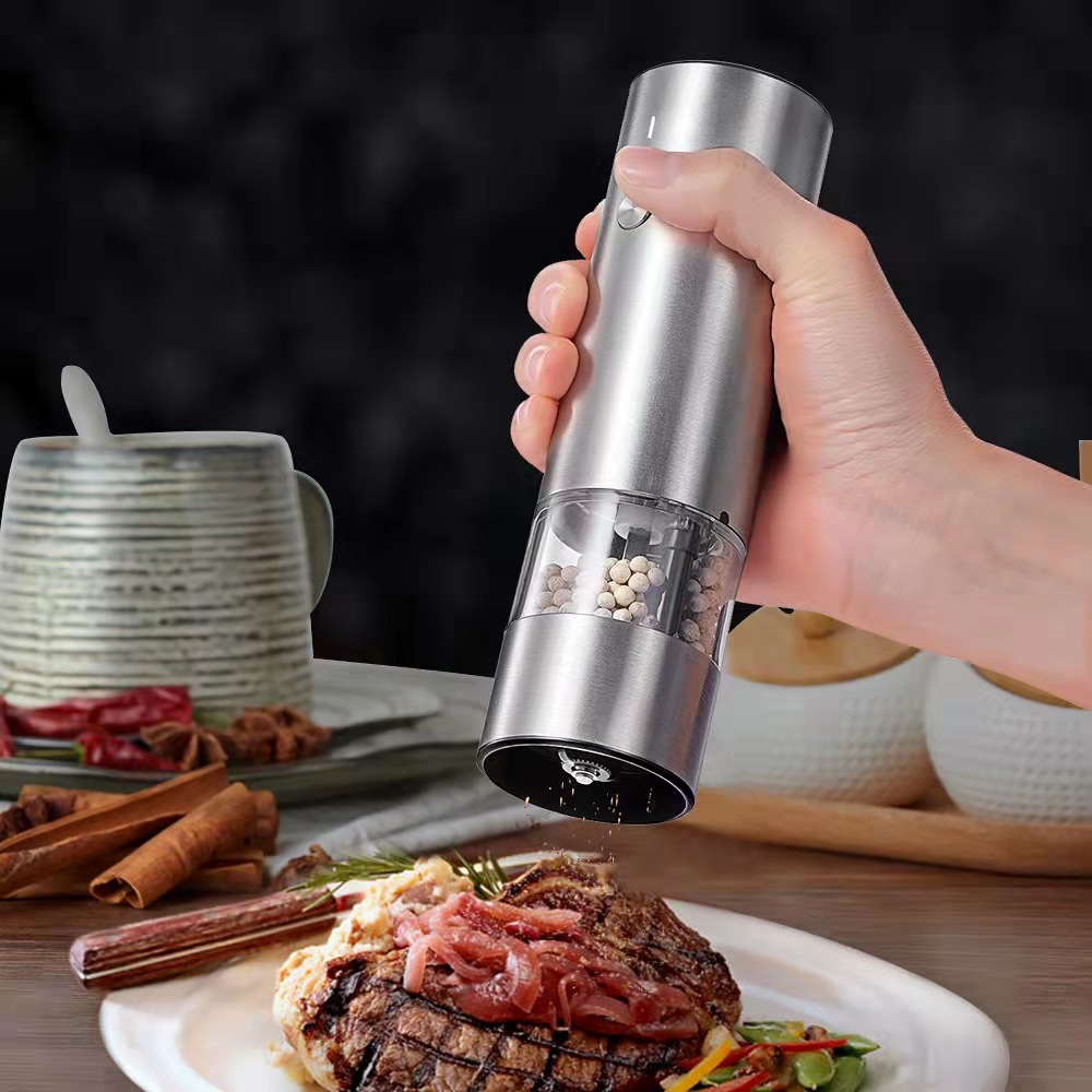 Eine Hand hält eine silberne elektrische Pfeffermühle und mahlt Pfeffer auf ein Steak auf einem weißen Teller. Im Hintergrund Gewürze und ein Becher.