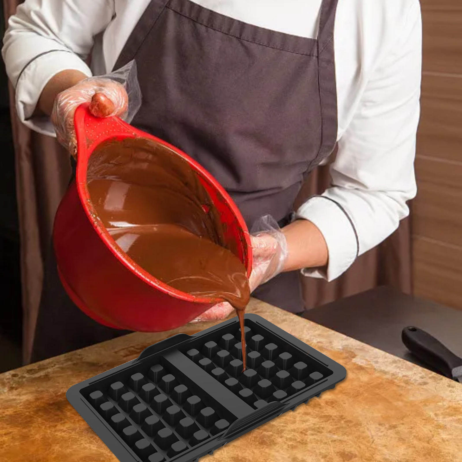 Ein Koch gießt Schokoladenteig aus einer roten Schüssel in ein schwarzes Waffeleisen. Die Szene spielt auf einer Holzoberfläche.