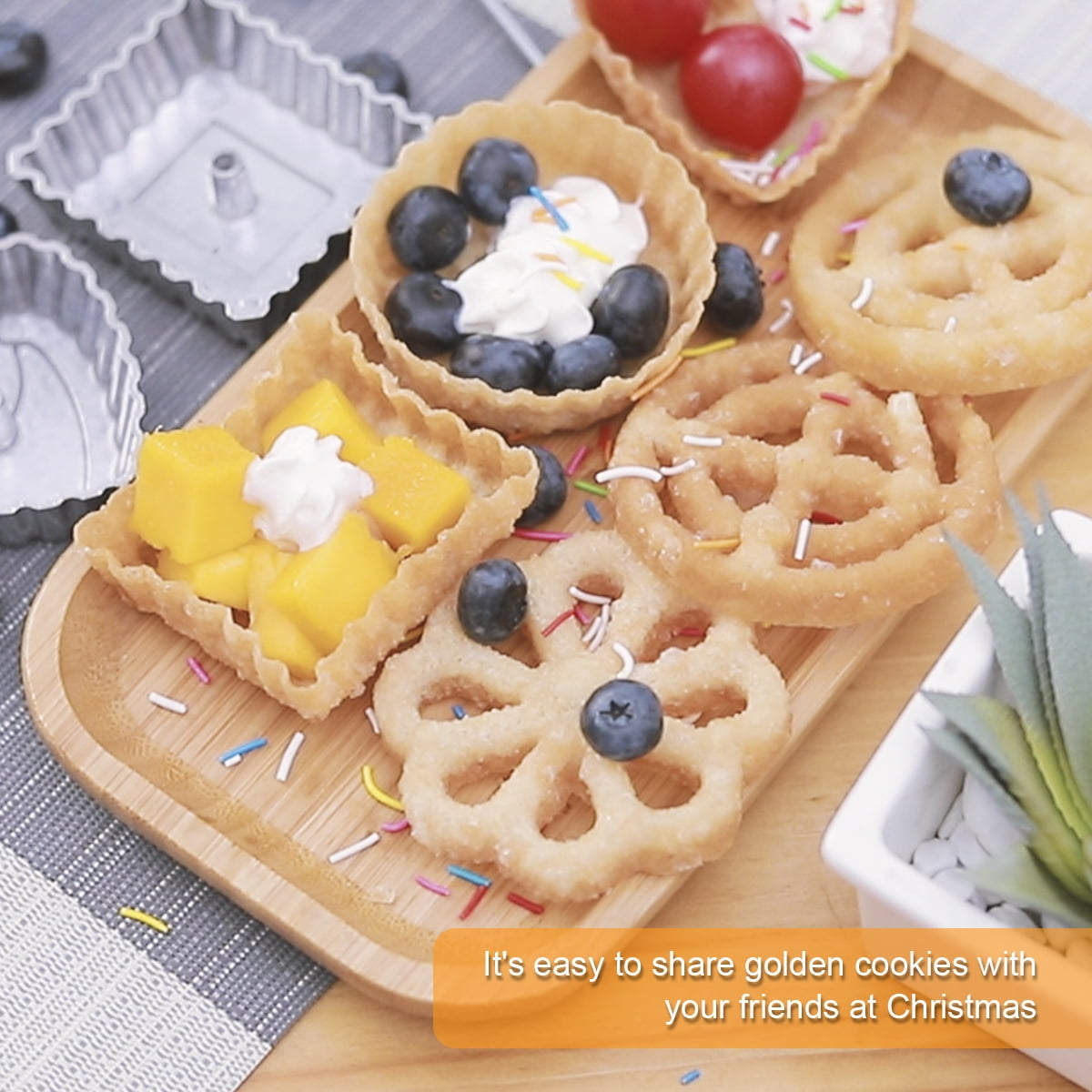 Holztablett mit Keksen, Früchten und Sahne. Einige Kekse sind blumenförmig. Blaubeeren und Mangos sind sichtbar.