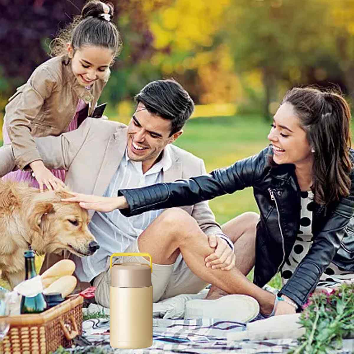 Familienpicknick mit Hund. Menschen streicheln den Hund. Ein Picknickkorb und eine Thermoskanne sind im Vordergrund. Der Hintergrund ist eine grüne Wiese.
