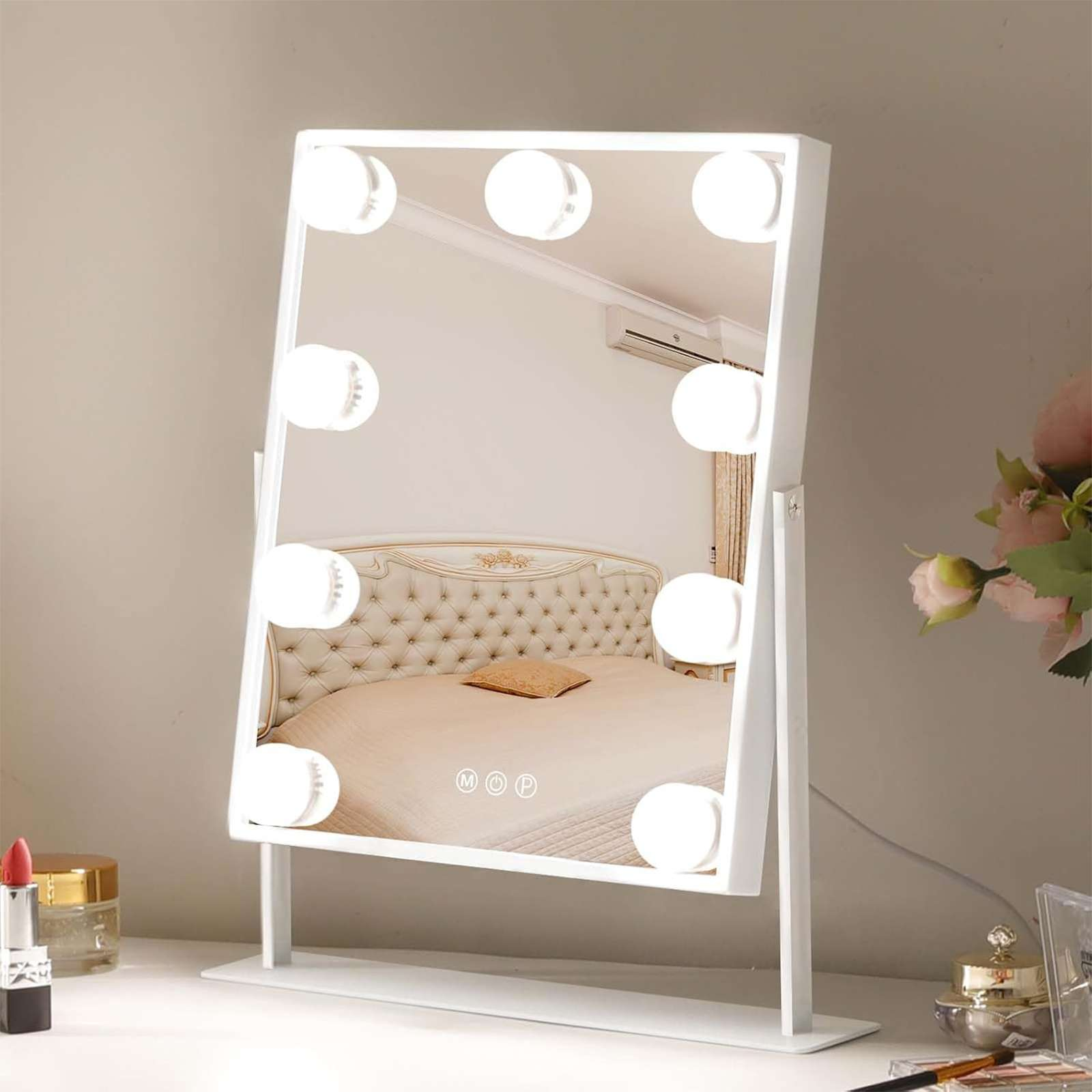 Weißer Schminkspiegel mit Lichtern. Die Spiegelung zeigt ein Bett, Blumen und Schminkartikel.