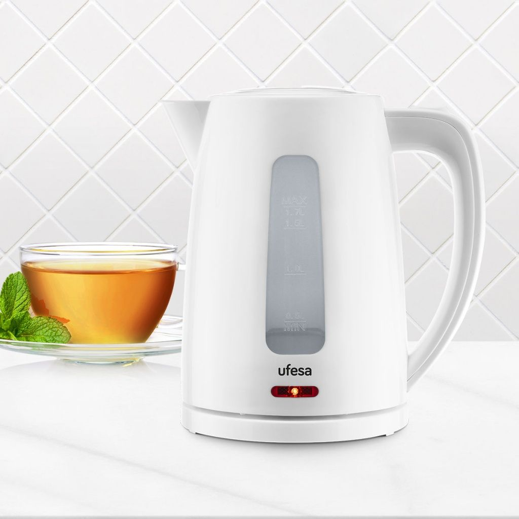 Un bollitore elettrico bianco con indicatore livello acqua trasparente e una tazza di tè con foglie di menta su superficie bianca.