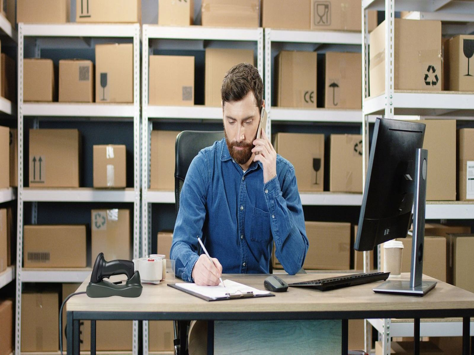 Mężczyzna w magazynie przez telefon, piszący. Pudełka na półkach w tle. Komputer, skaner i kubek kawy na biurku.