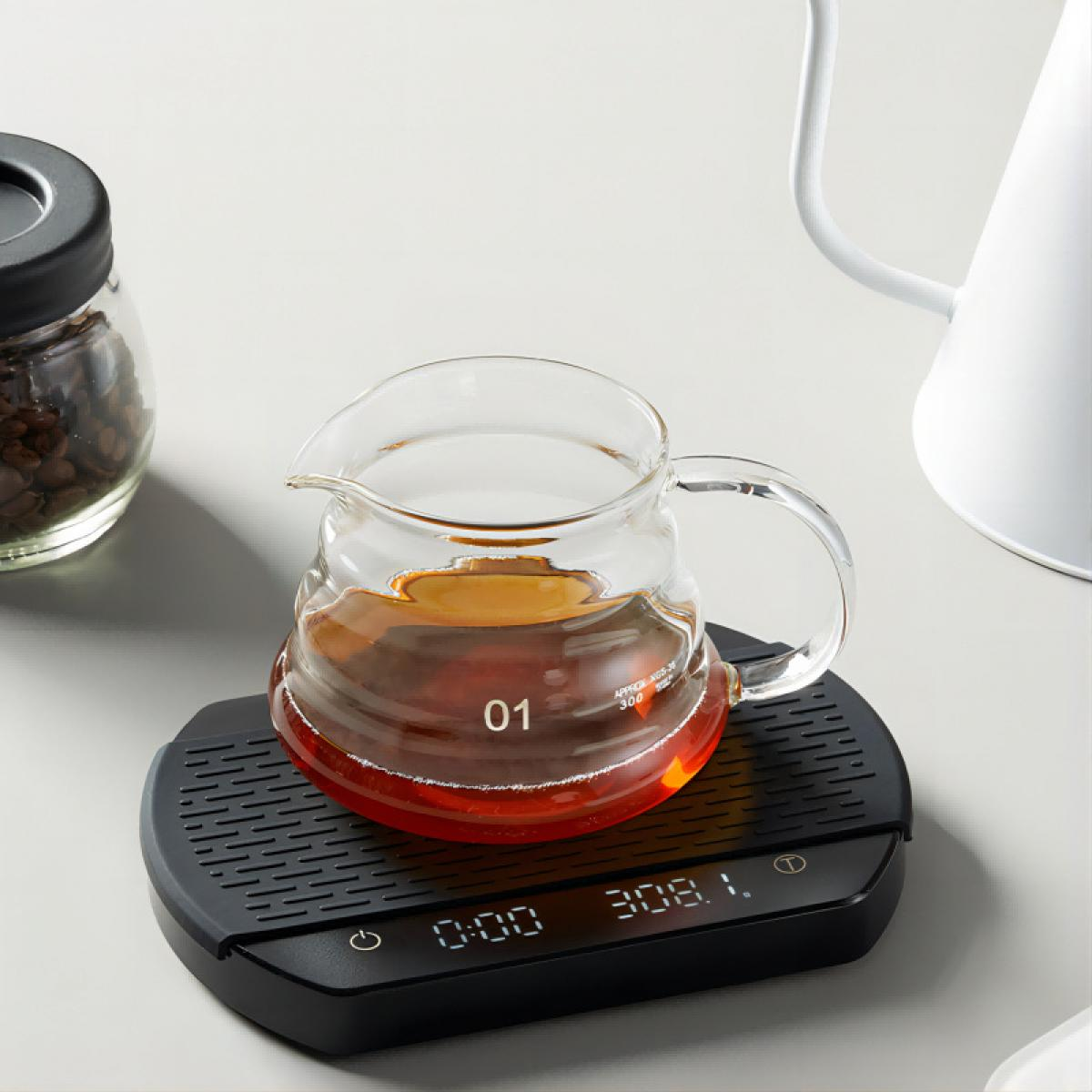 Eine Glaskaraffe mit Kaffee steht auf einer schwarzen Waage. Ein Glas Kaffeebohnen und ein weißer Wasserkocher sind ebenfalls vorhanden.