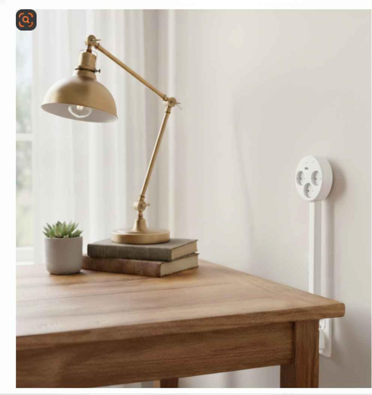 Ein Schreibtisch mit Lampe, Büchern und Pflanze. Eine Steckdose ist an der Wand. Die Lampe hat eine goldene Oberfläche.