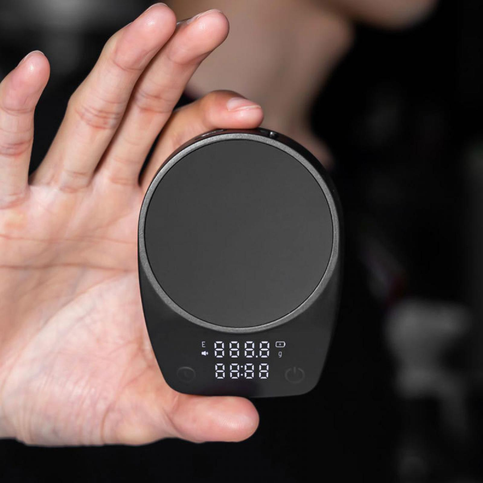 Eine Hand hält eine schwarze Digitalwaage. Das Display zeigt Zahlen. Der Hintergrund ist verschwommen.