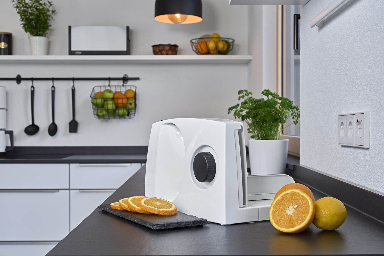 Scena kuchenna z białą krajalnicą, plastrami pomarańczy i cytryn na czarnym blacie. Widoczne owoce i rośliny.