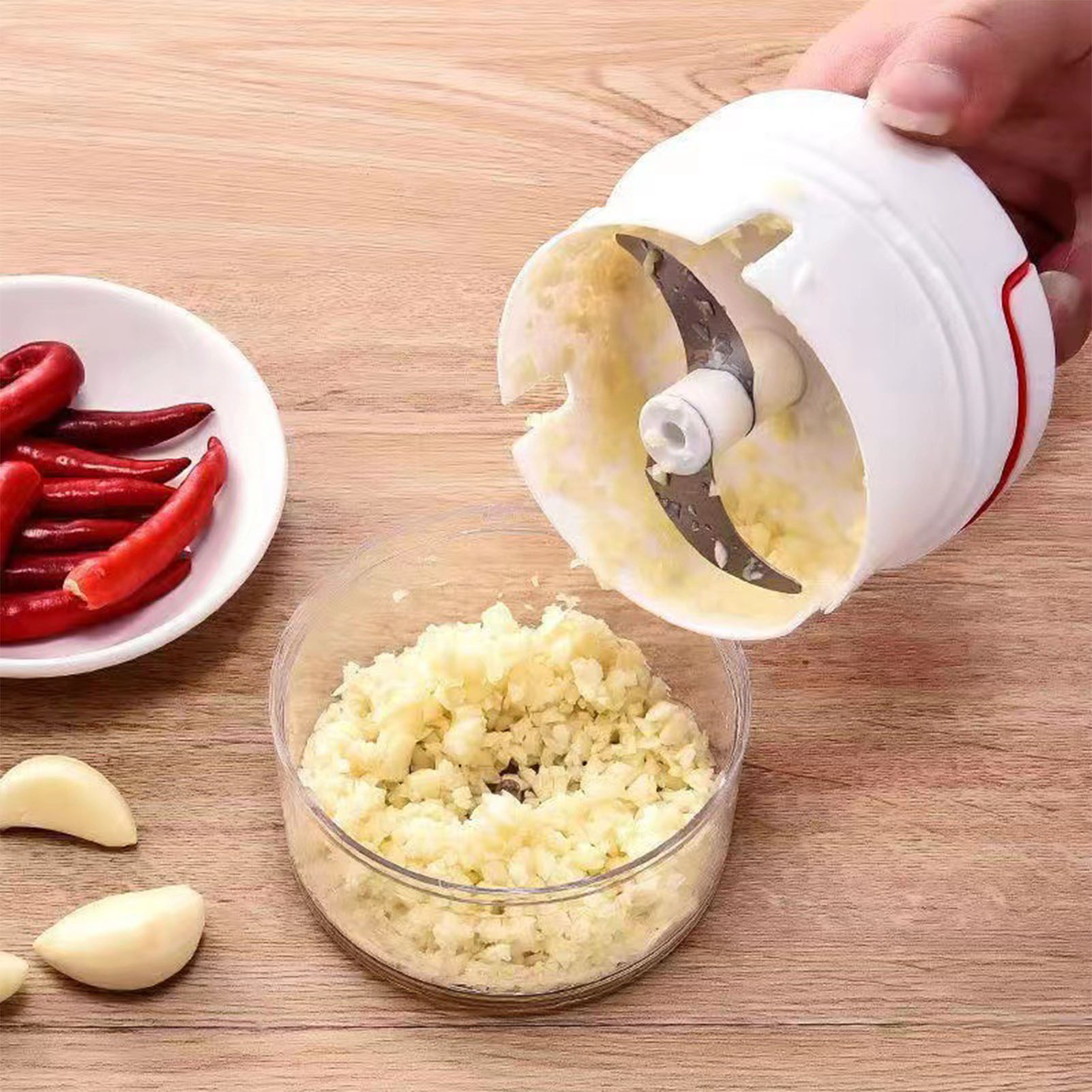 Ein weißer Knoblauchschneider ist geöffnet, Knoblauch wird gehackt. Rote Paprika und Knoblauchzehen liegen neben dem Schneider.