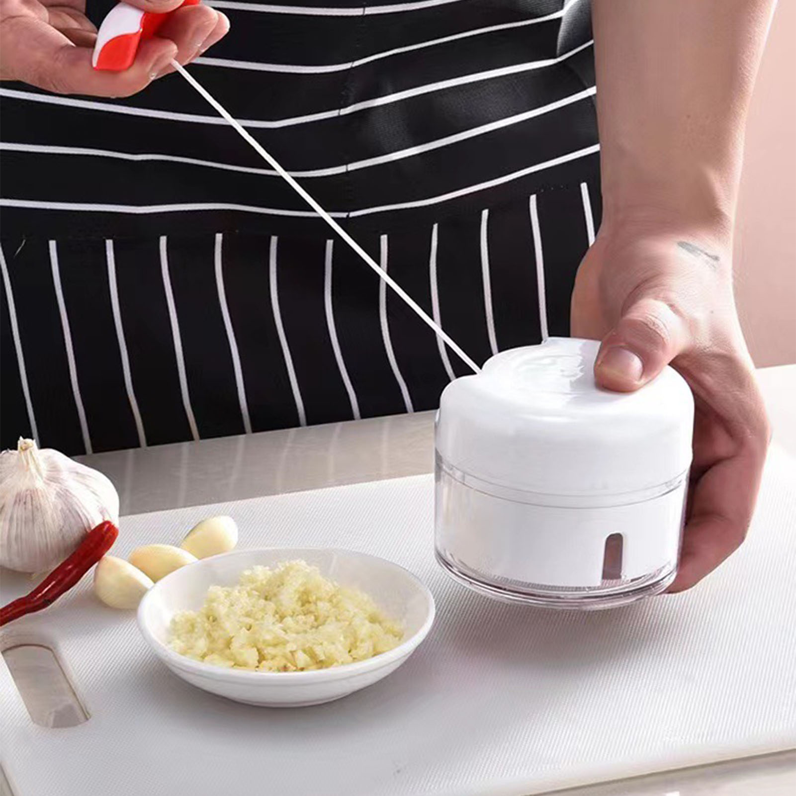 Eine Person benutzt einen Knoblauchschneider. Zutaten sind Knoblauch, eine rote Paprika und gehackter Knoblauch in einer Schüssel auf einem weißen Brett.