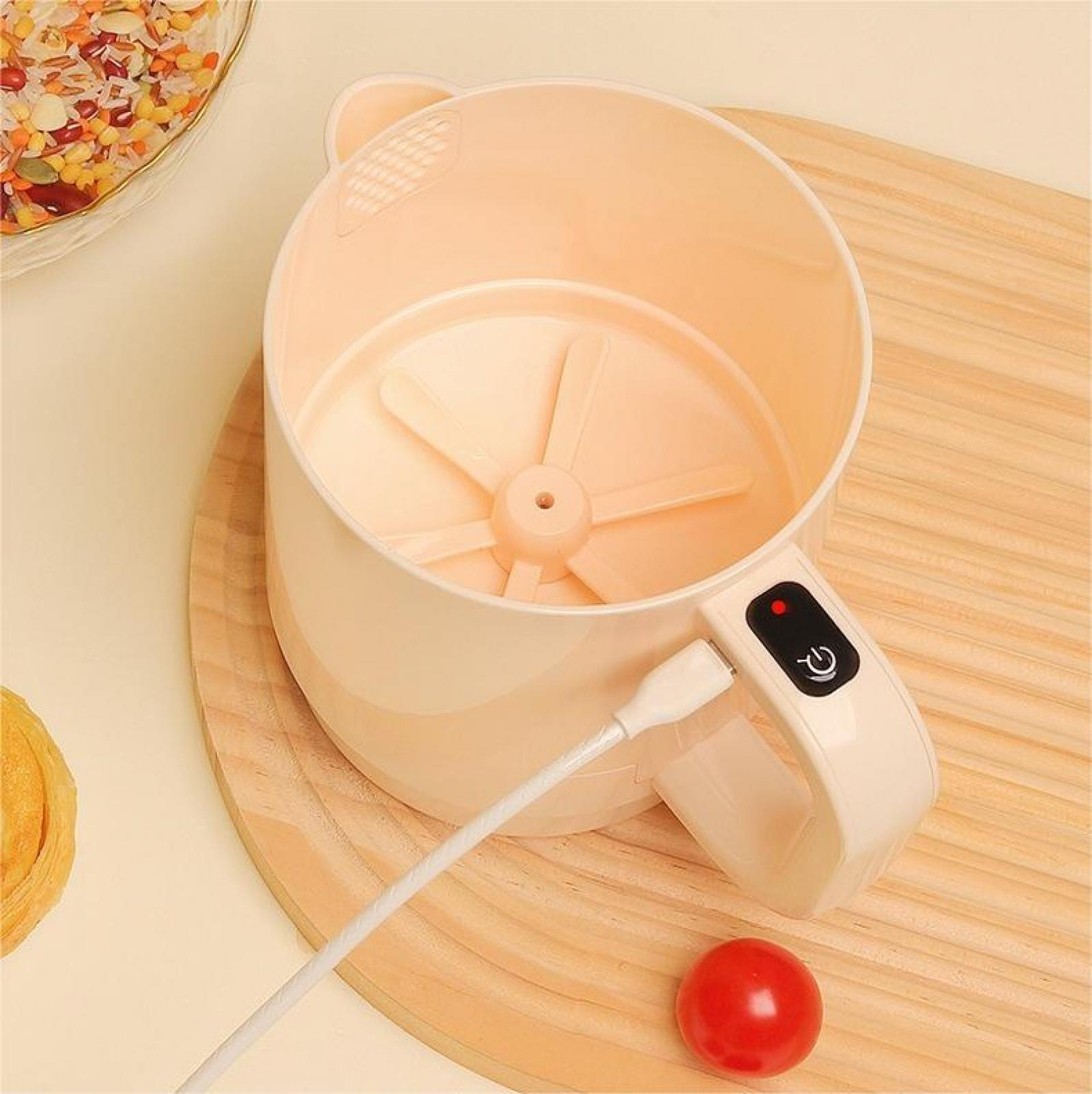 Ein beiger Mixer mit weißem USB-Kabel und einer roten Kirsche steht auf einem Holzbrett. Eine Schüssel mit Samen ist im Hintergrund.