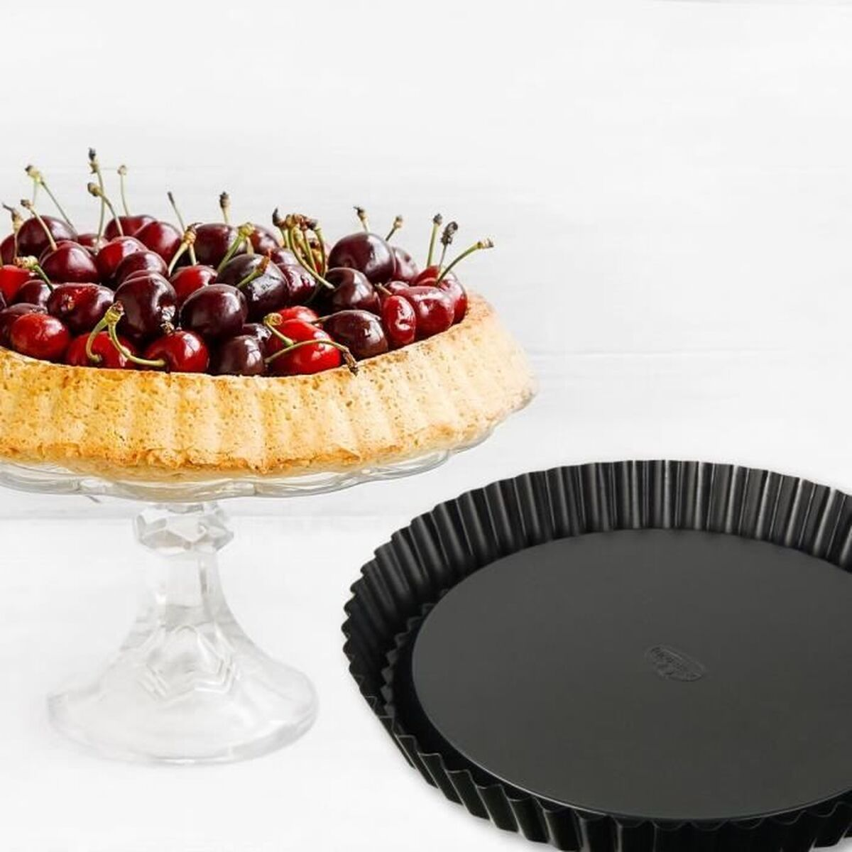 Kirschkuchen auf einem Glasständer und eine schwarze Tortenform auf weißem Untergrund. Der Kuchen ist mit Kirschen gefüllt.