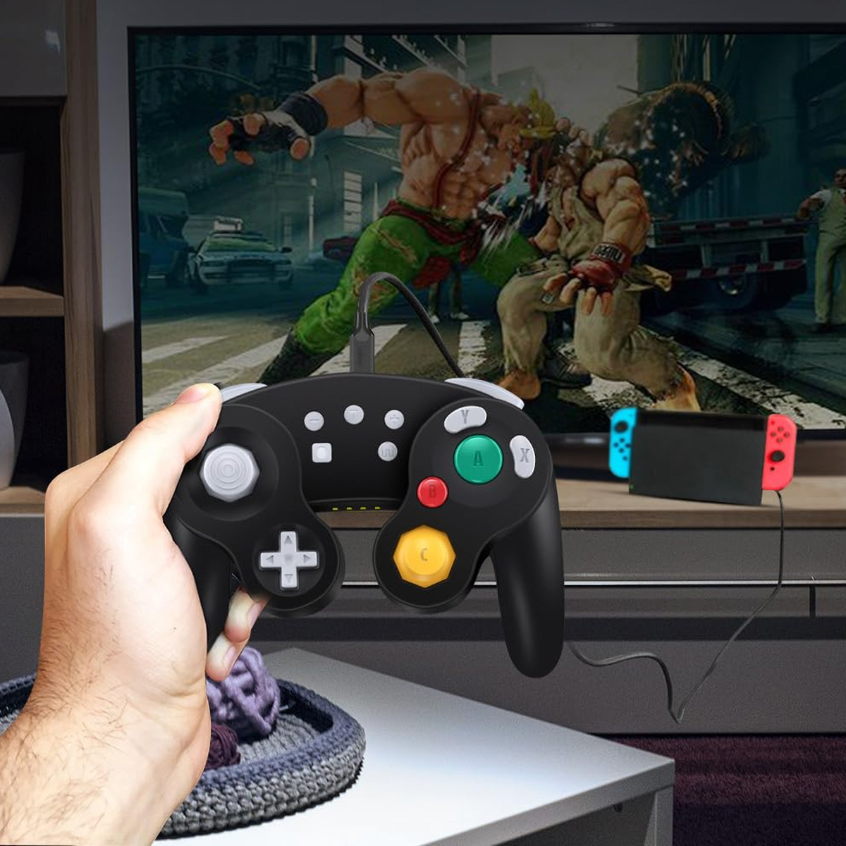 Eine Hand hält einen schwarzen Gamecontroller. Ein Fernseher zeigt ein Kampfspiel. Rechts ist eine Nintendo Switch. Links ein Korb.